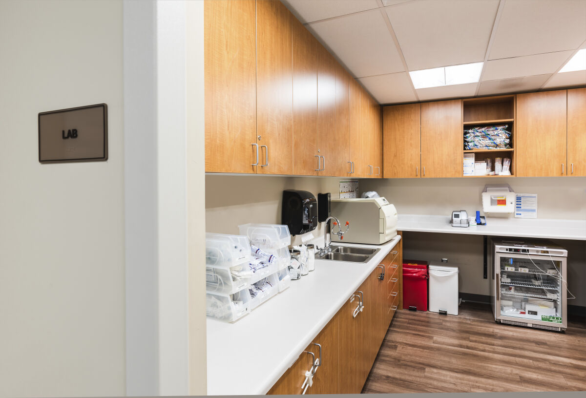 Lab room with wood cabinetry, solid-surface countertops, a stainless-steel sink, storage bins with medical supplies, diagnostic equipment, and wood-look flooring under bright ceiling lights.
