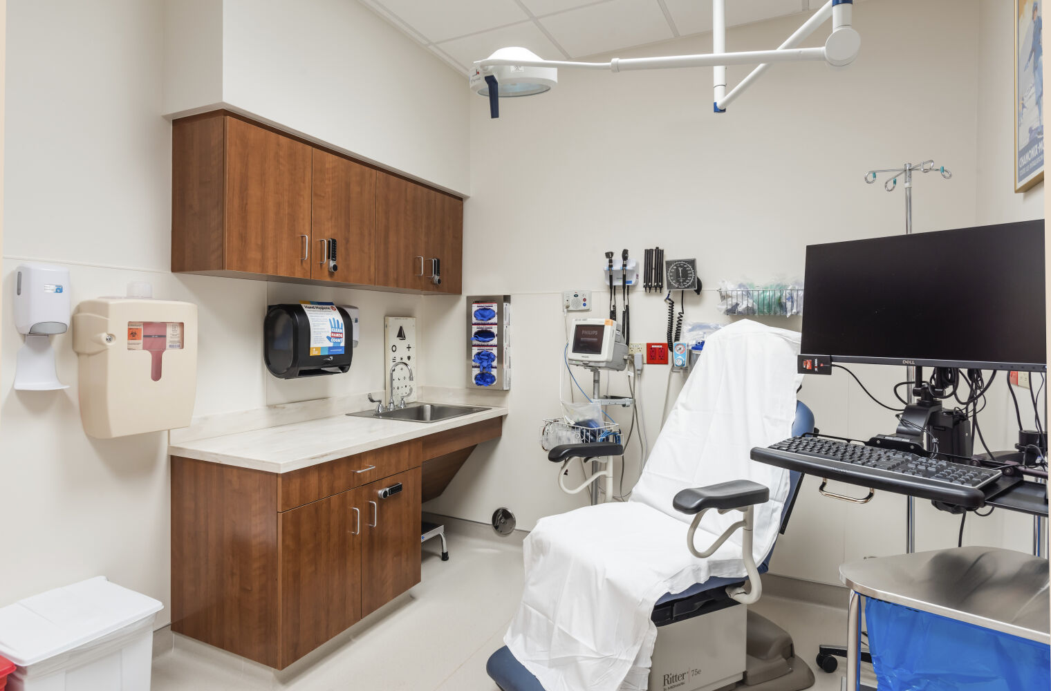 Exam room with a reclining exam chair covered in a white sheet, wall-mounted diagnostic equipment, wood cabinetry with a sink, ceiling-mounted procedure light, IV pole, and a computer workstation on an adjustable arm.