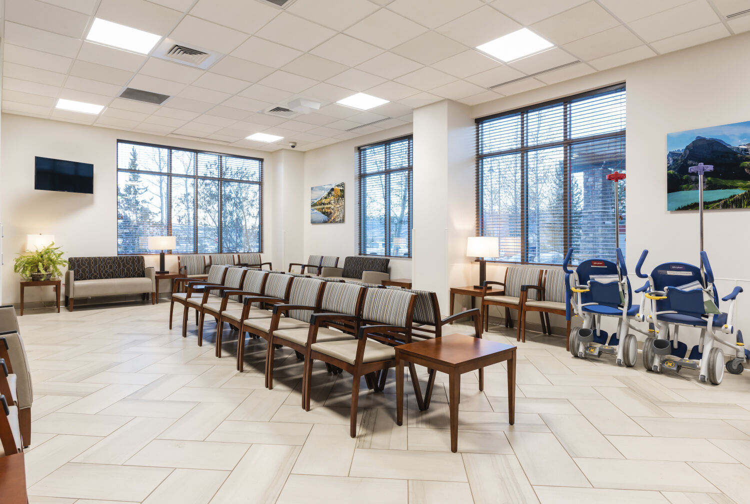 Emergency department waiting area with rows of upholstered chairs, wood side tables, wall-mounted landscape artwork, a water dispenser, large windows, tile flooring, and a view toward the reception desk and staff corridor.