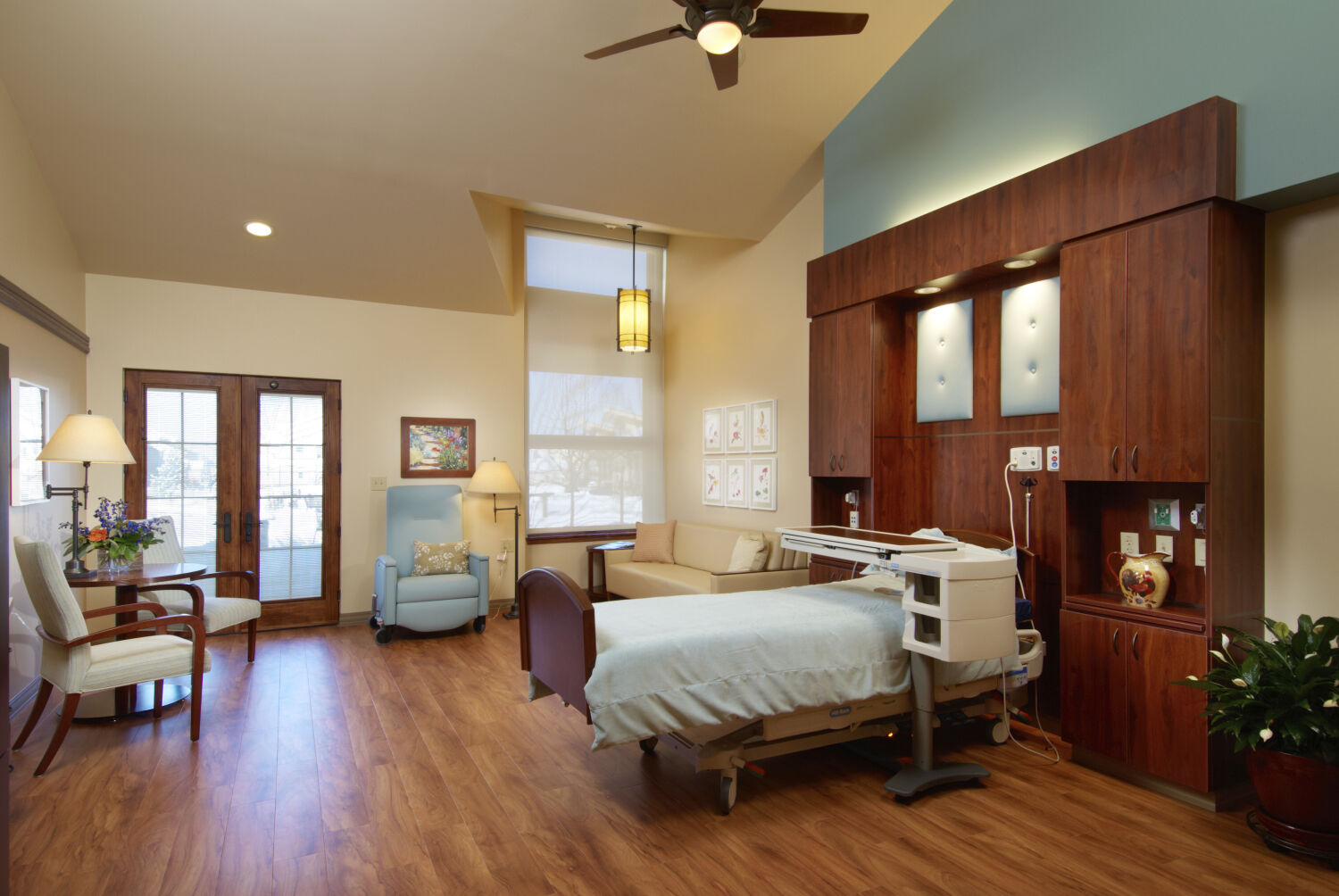 Patient room with a hospital bed, wood cabinetry and headwall unit, upholstered recliner, sofa, wood armchairs, bedside lighting, framed artwork, wood-look flooring, and double glass doors leading to an outdoor patio.
