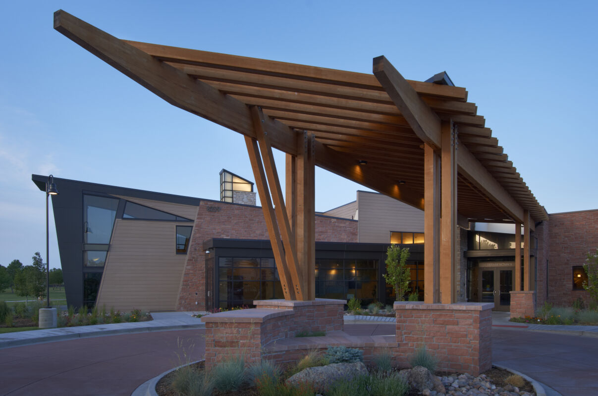 Exterior entrance featuring a large timber canopy structure with angled supports, brick seating walls, landscaped planting beds, and a modern combination of brick, glass, and metal façade elements.