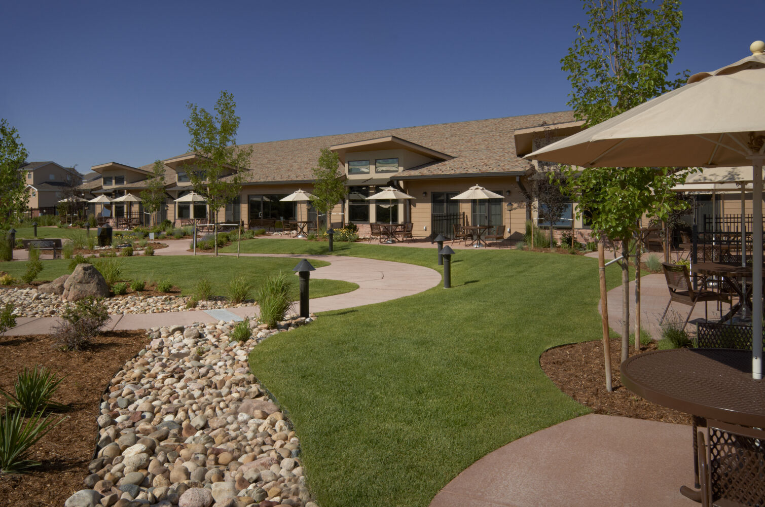 Garden courtyard at The Denver Hospice with curved walkways, landscaped planting beds with rocks and shrubs, lawn areas, patio tables with umbrellas, and exterior building façades with large windows and covered seating areas.