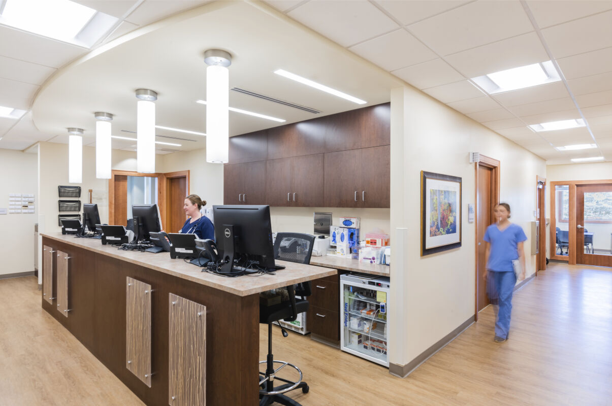 Nurse station with a long wood work counter featuring multiple computer monitors, overhead pendant lighting, upper wood cabinets, a compact undercounter refrigerator with medical supplies, and adjacent wood doors leading to treatment and support rooms.