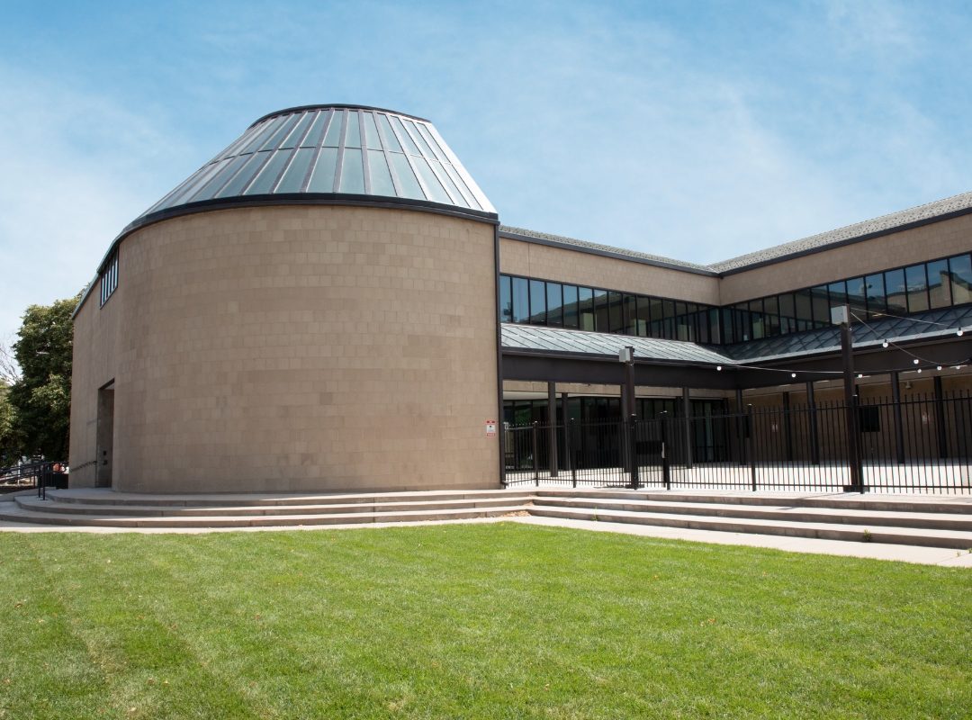 The Community College of Denver Clear Creek Building featuring a large cylindrical volume with a sloped glass roof, linear wings with continuous ribbon windows, a covered walkway with metal columns, concrete steps, and an open lawn in the foreground.