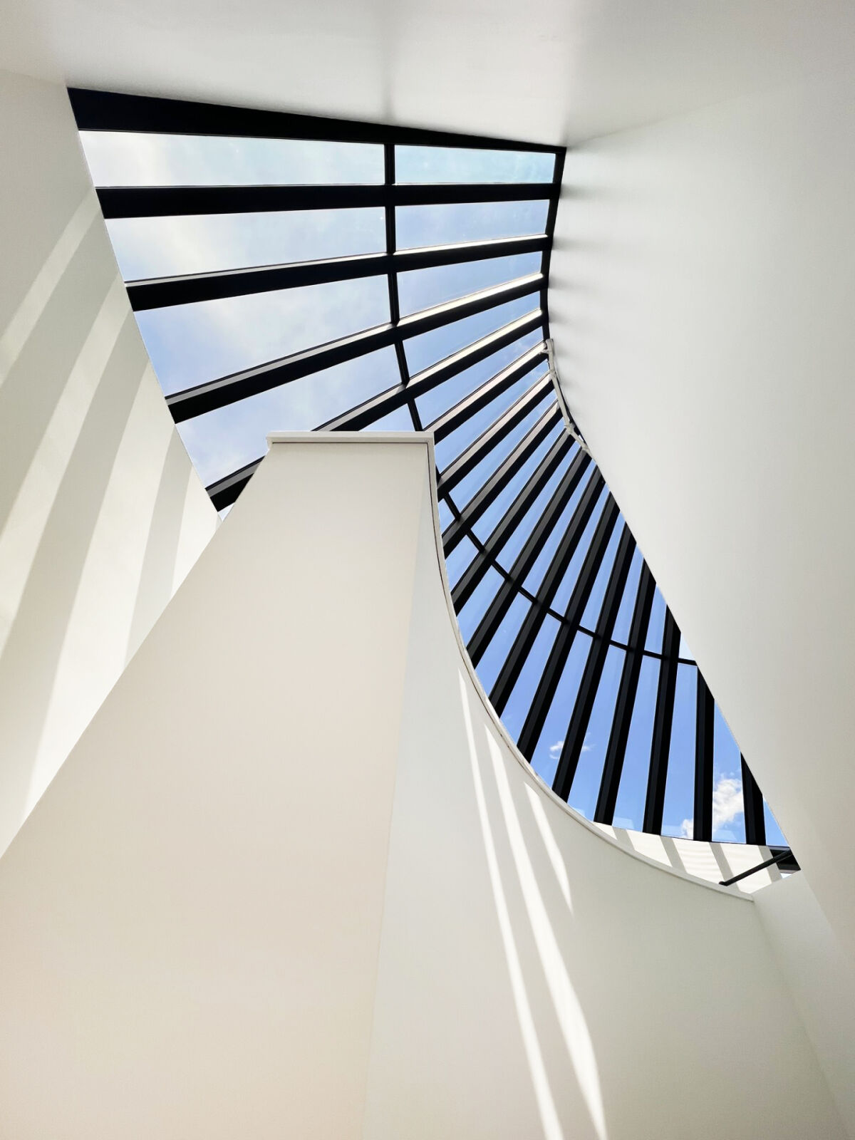Upward view of a curved interior wall and large skylight with black framing, casting angled daylight shadows.
