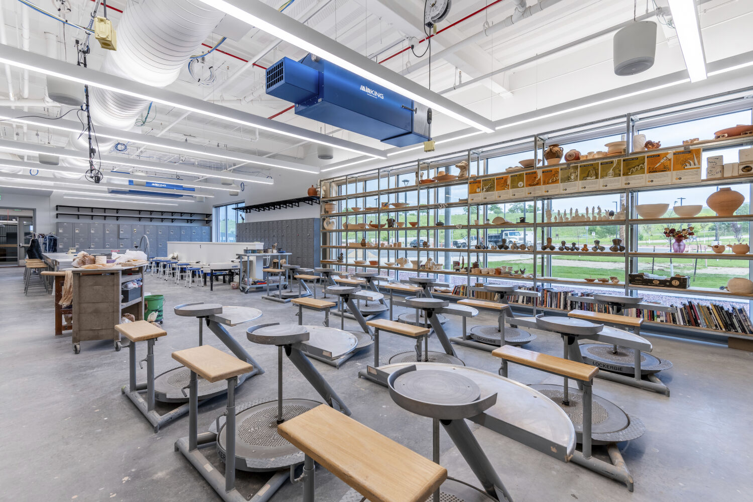 Ceramics studio in the Casper College Tobin Visual Arts Center with rows of pottery wheels, long worktables, extensive shelving filled with ceramic pieces and supplies, overhead ductwork, and polished concrete flooring.