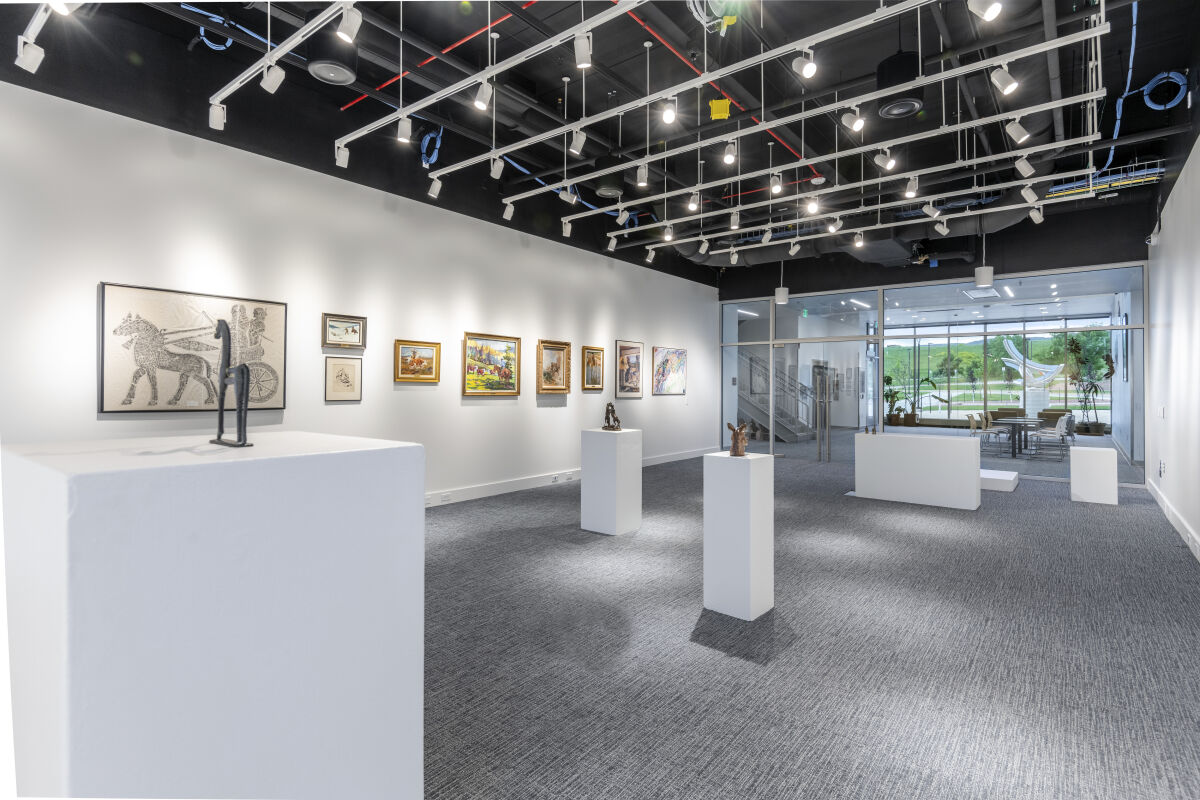 Gallery in the Casper College Tobin Visual Arts Center with artwork displayed on white walls, sculptures on white pedestals, carpeted flooring, suspended track lighting, an open ceiling grid, and glass doors leading to an adjacent lobby area.