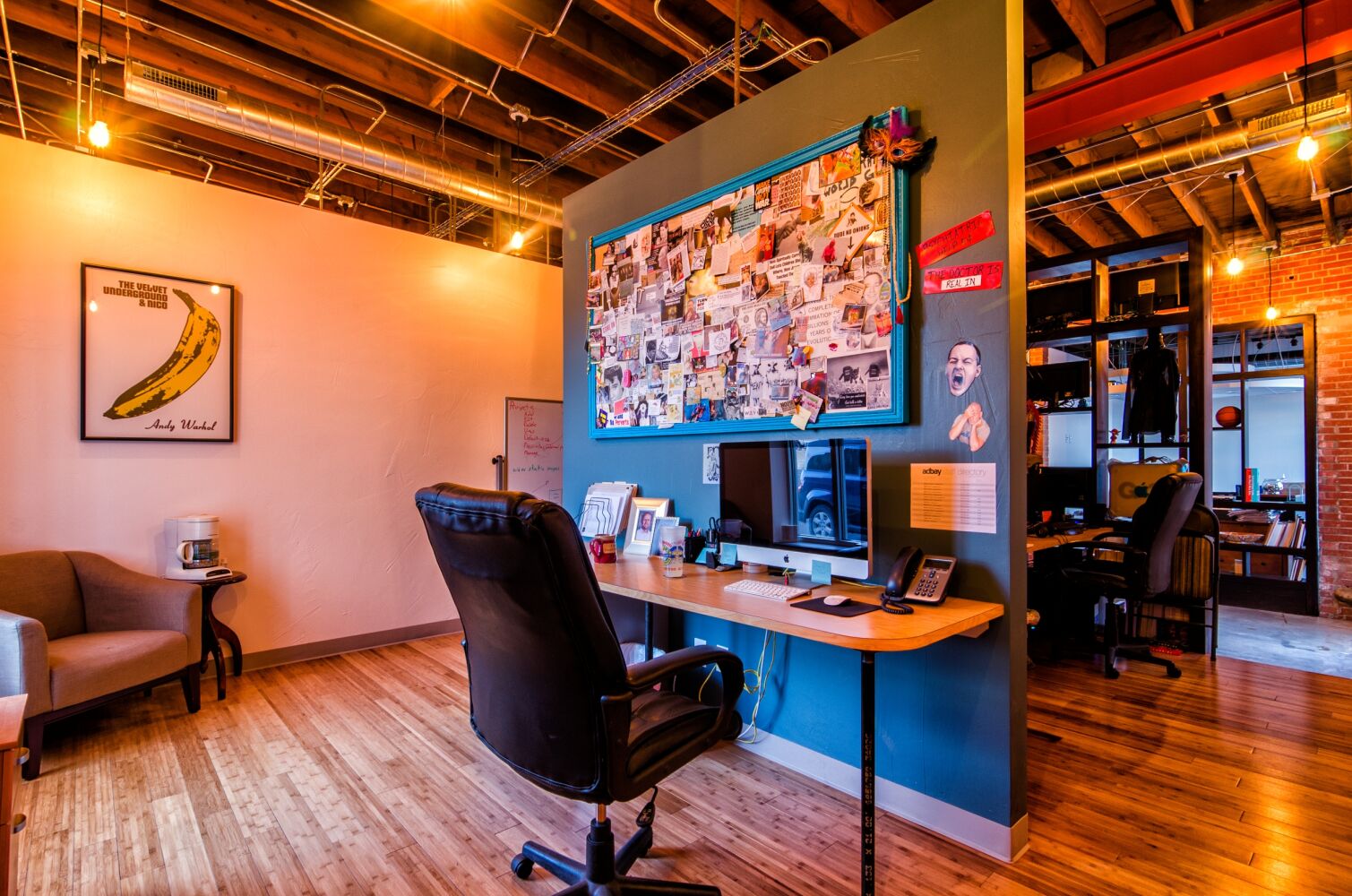 Creative office workspace with exposed wood ceilings and ductwork, a desk with computer monitors, a large photo collage on a blue accent wall, wood flooring, and an adjacent lounge chair beneath artwork.