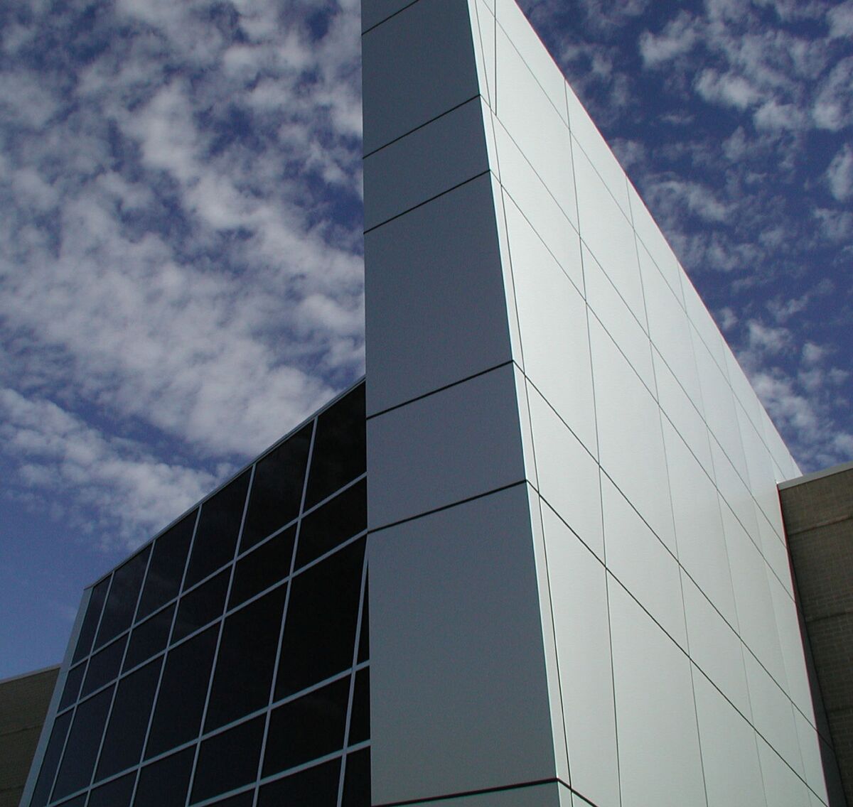 Close‑up exterior view of an angular glass and metal façade with grid‑pattern windows and smooth silver wall panels against a sky with scattered clouds.