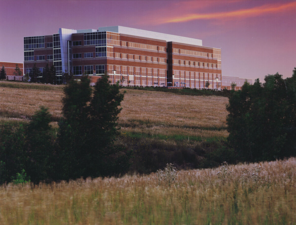 Distant view of a multi‑story office building with brick and glass façades, set behind an open field with tall grasses and silhouetted trees at sunset.
