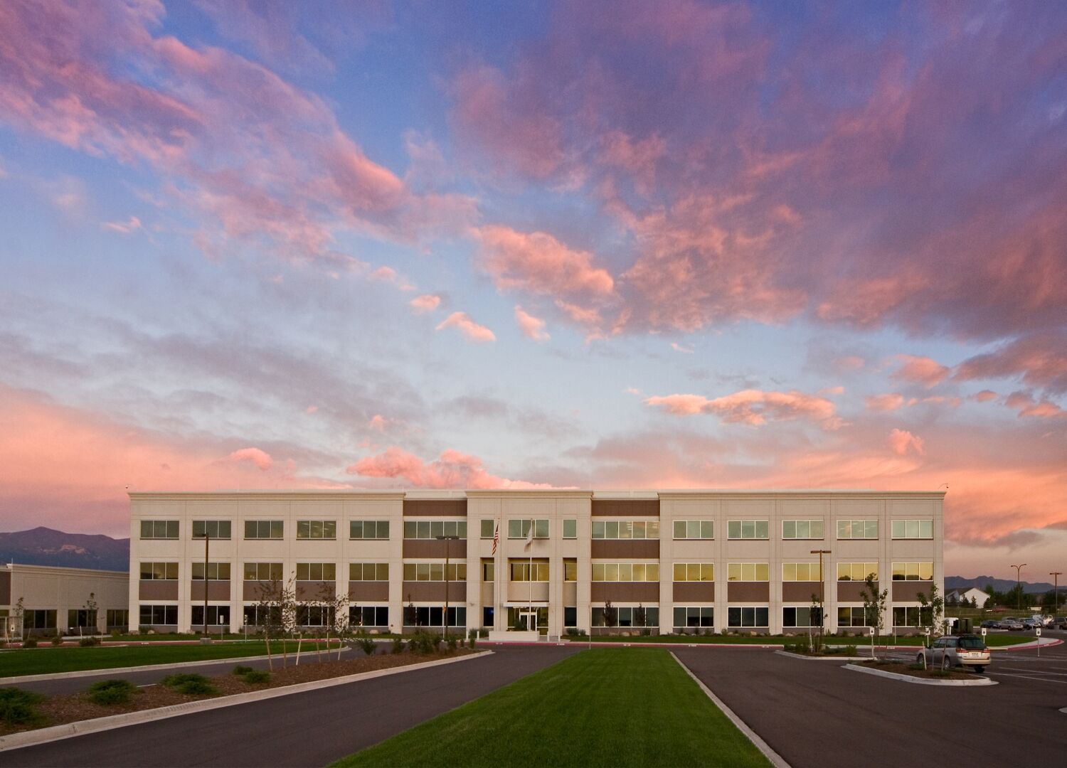 Exterior view of a three‑story office building with large windows, symmetrical façade, landscaped grounds, and a central walkway, shown at sunset beneath colorful clouds.