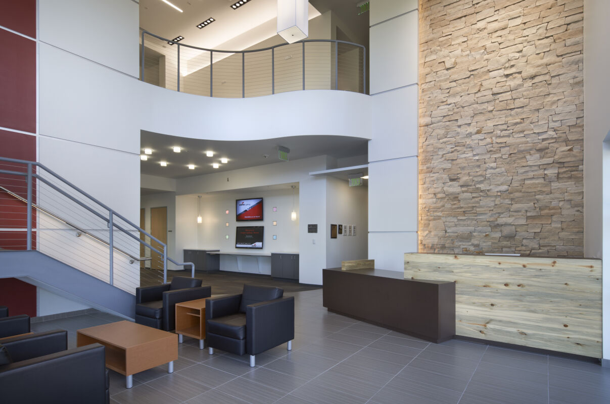 Modern lobby interior with a double‑height space, stone feature wall, wood reception desk, metal stair railing, and seating area with lounge chairs.