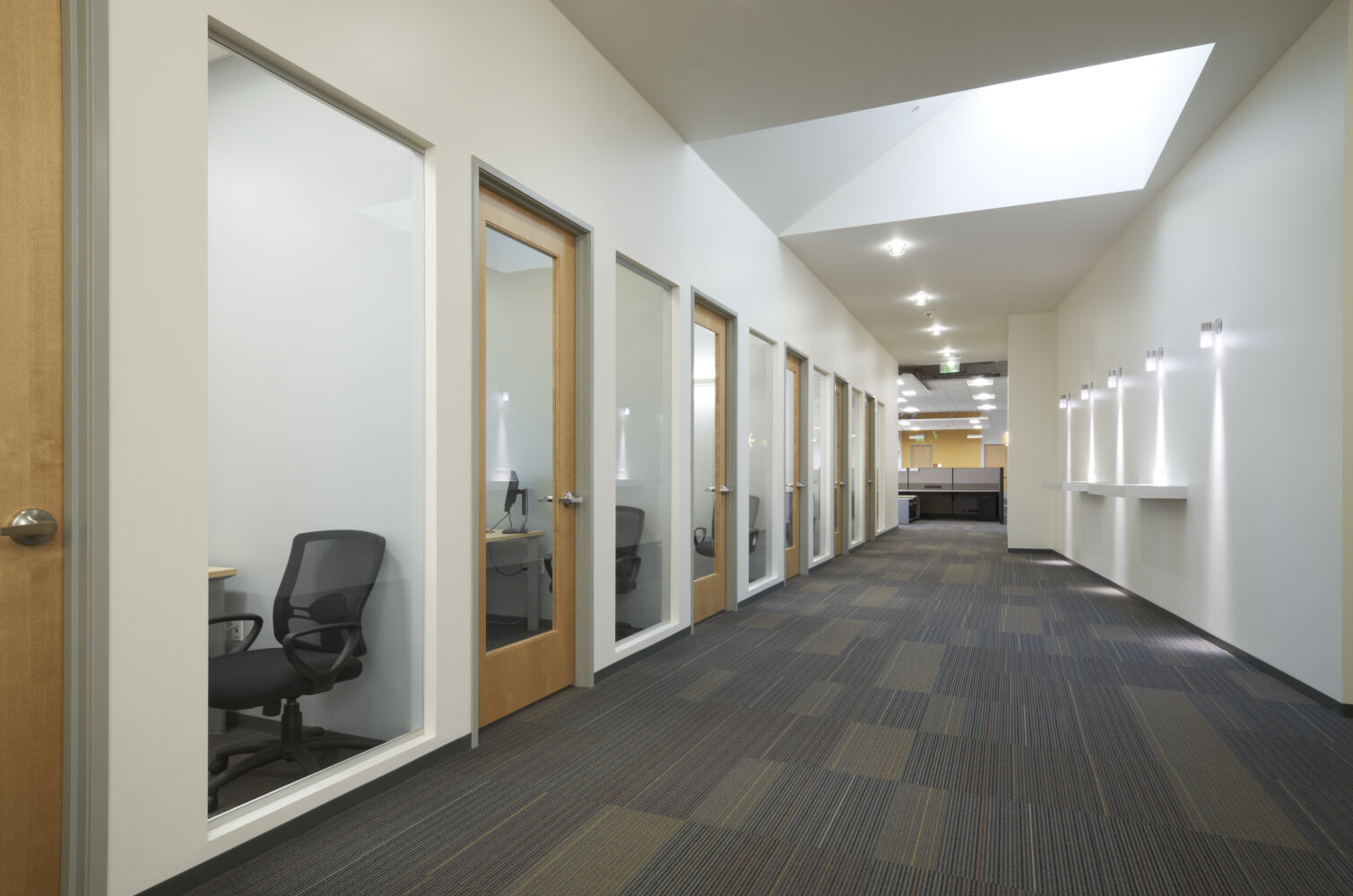 Office corridor with a row of private rooms featuring glass‑paneled doors, carpet tile flooring, recessed ceiling lighting, and a large skylight bringing in natural light.
