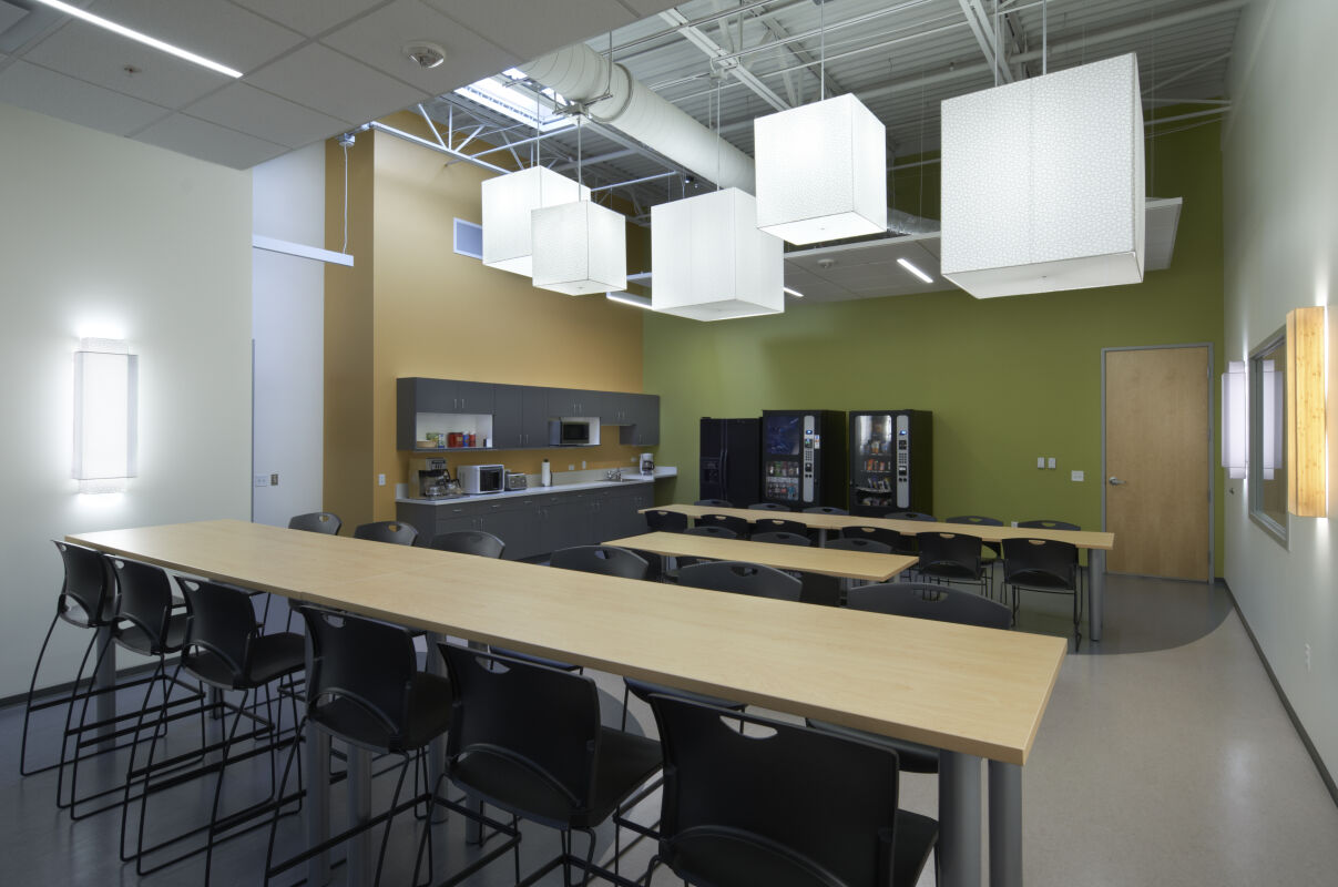 Breakroom with long high‑top tables, black chairs, suspended cube‑shaped light fixtures, a kitchenette with gray cabinets and appliances, and vending machines along a green accent wall.