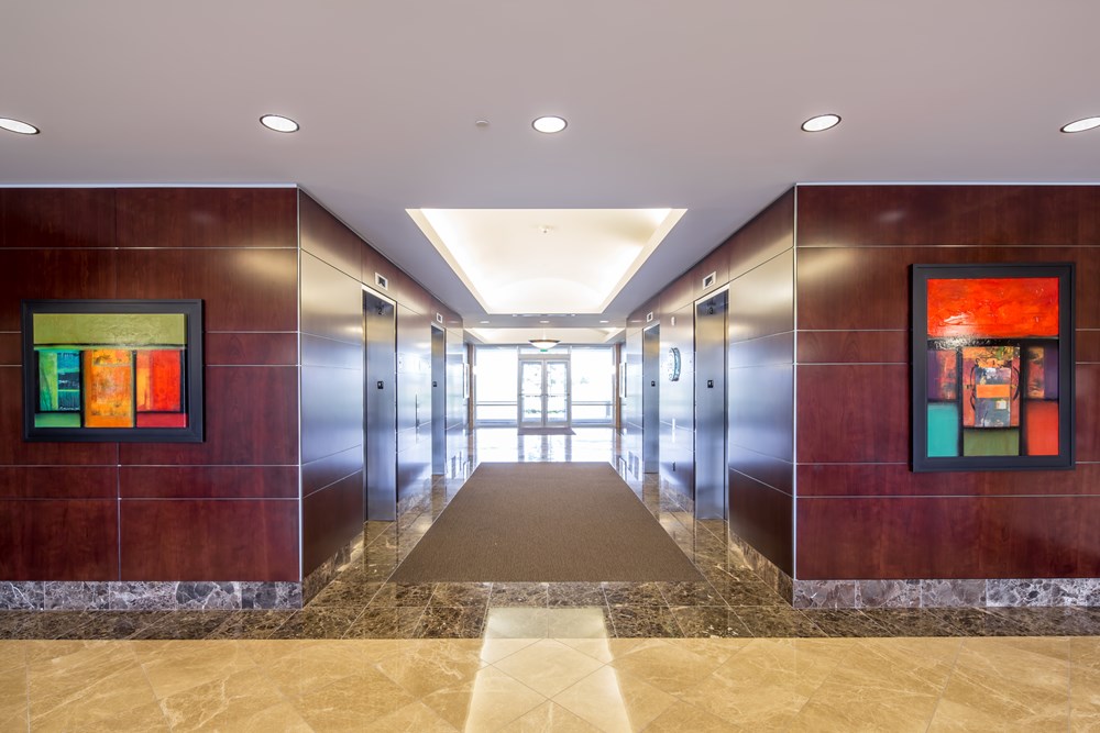 Elevator lobby with dark wood wall panels, marble flooring, recessed ceiling lighting, and colorful framed artwork flanking two sets of elevator doors.