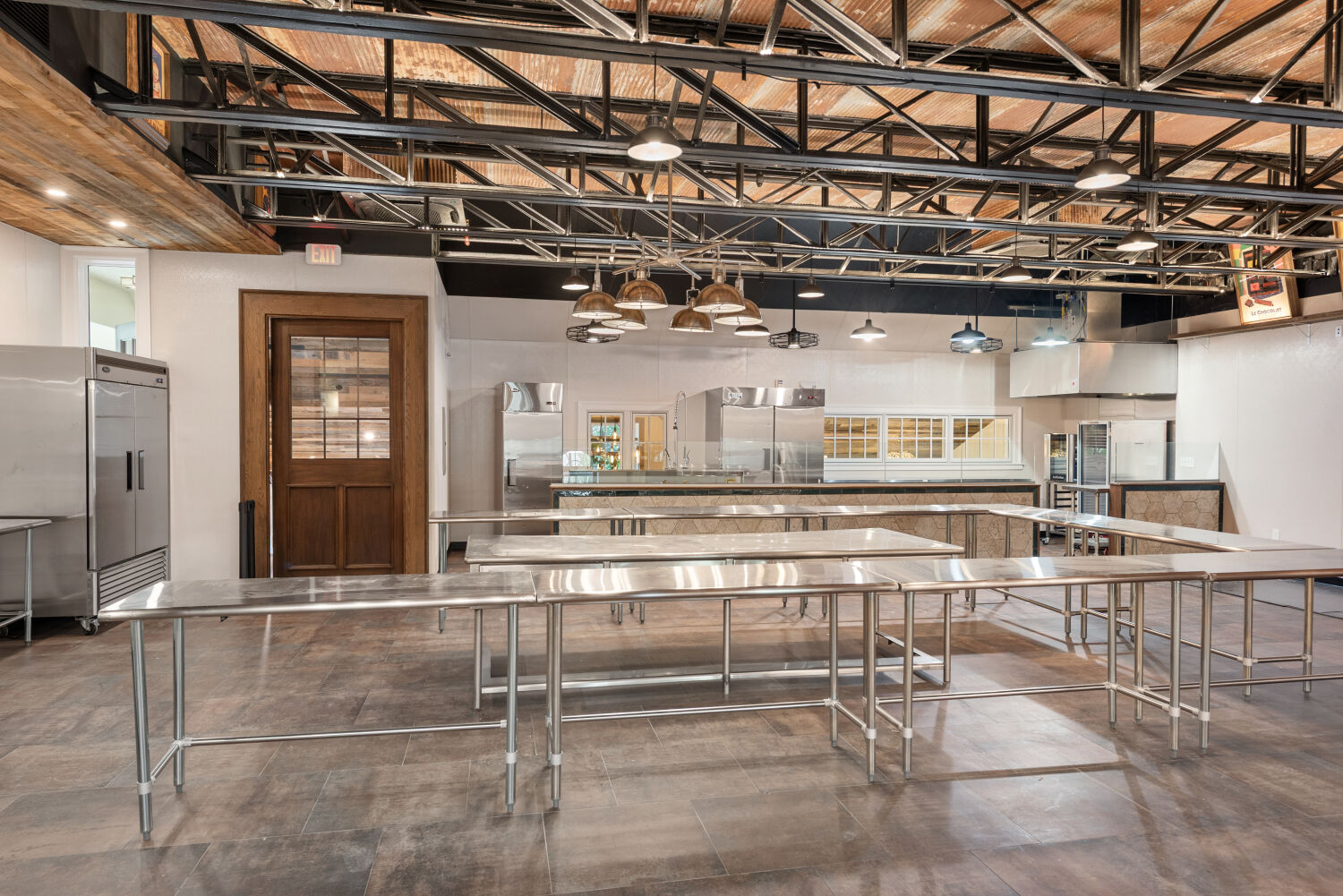 Commercial kitchen featuring stainless-steel prep tables, industrial appliances, exposed ceiling structure, and warm wood accents.