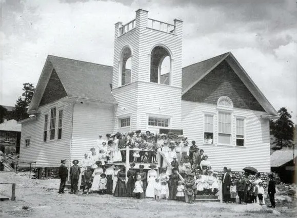 Historic black‑and‑white photograph of the Presbyterian Community Church of the Rockies, showing a large group of community members gathered on the front steps of the white wooden church with its central bell tower.
