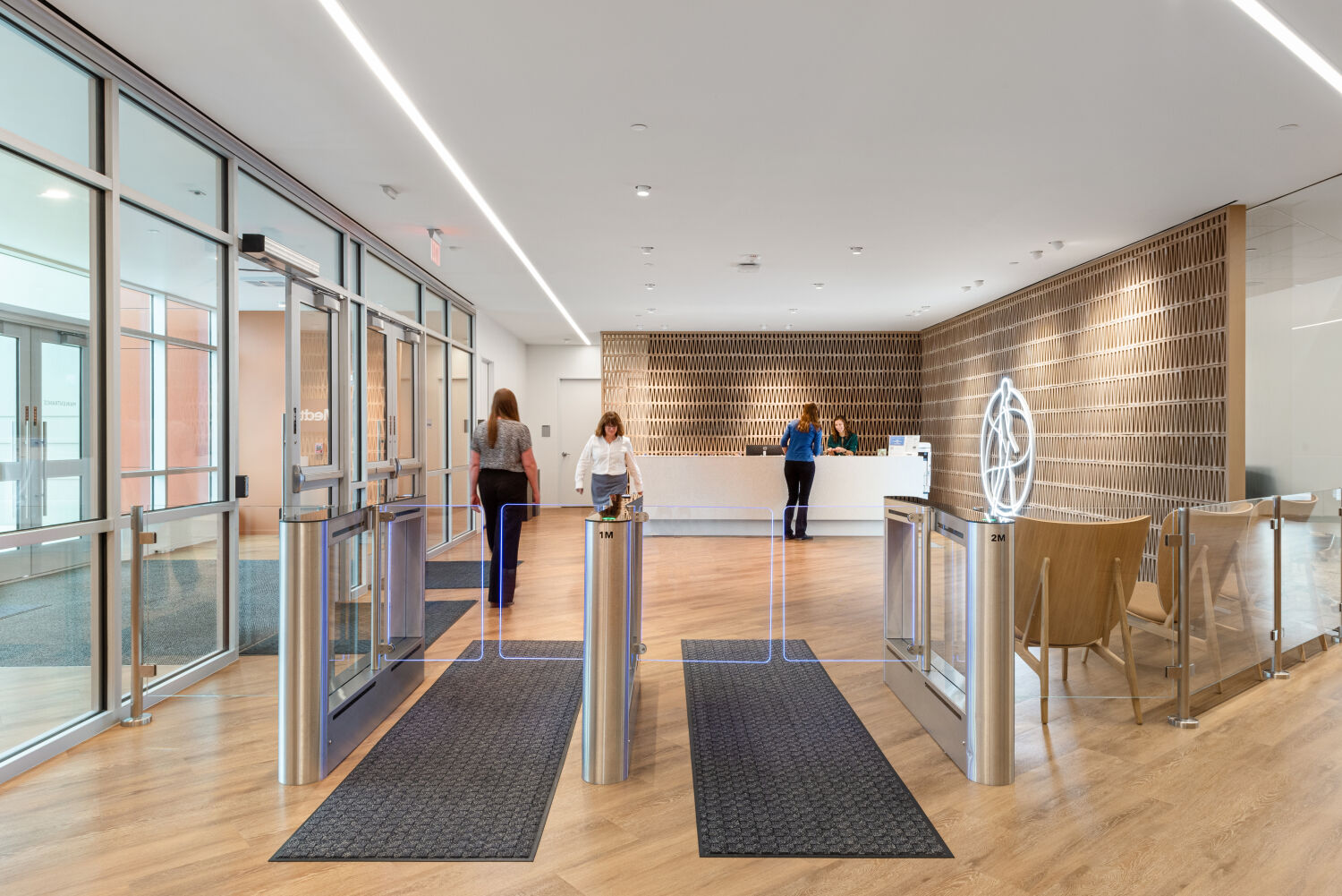 Reception lobby in a Medtronic commercial office featuring turnstile security gates, a wood‑accented reception desk, geometric wall paneling, glass entry doors, and overhead linear lighting.