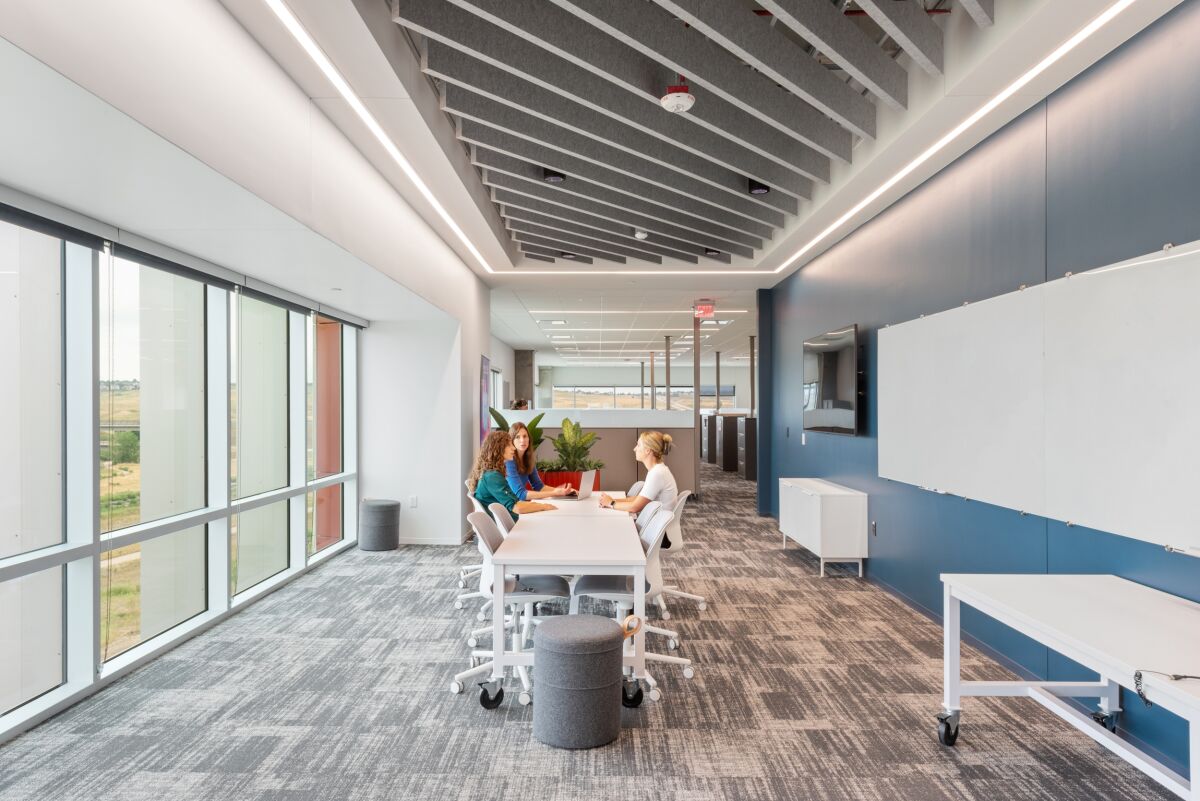 Bright open collaboration area in a Medtronic office featuring long tables with casters, acoustic ceiling baffles, large windows, gray patterned carpet, and writable wall panels.
