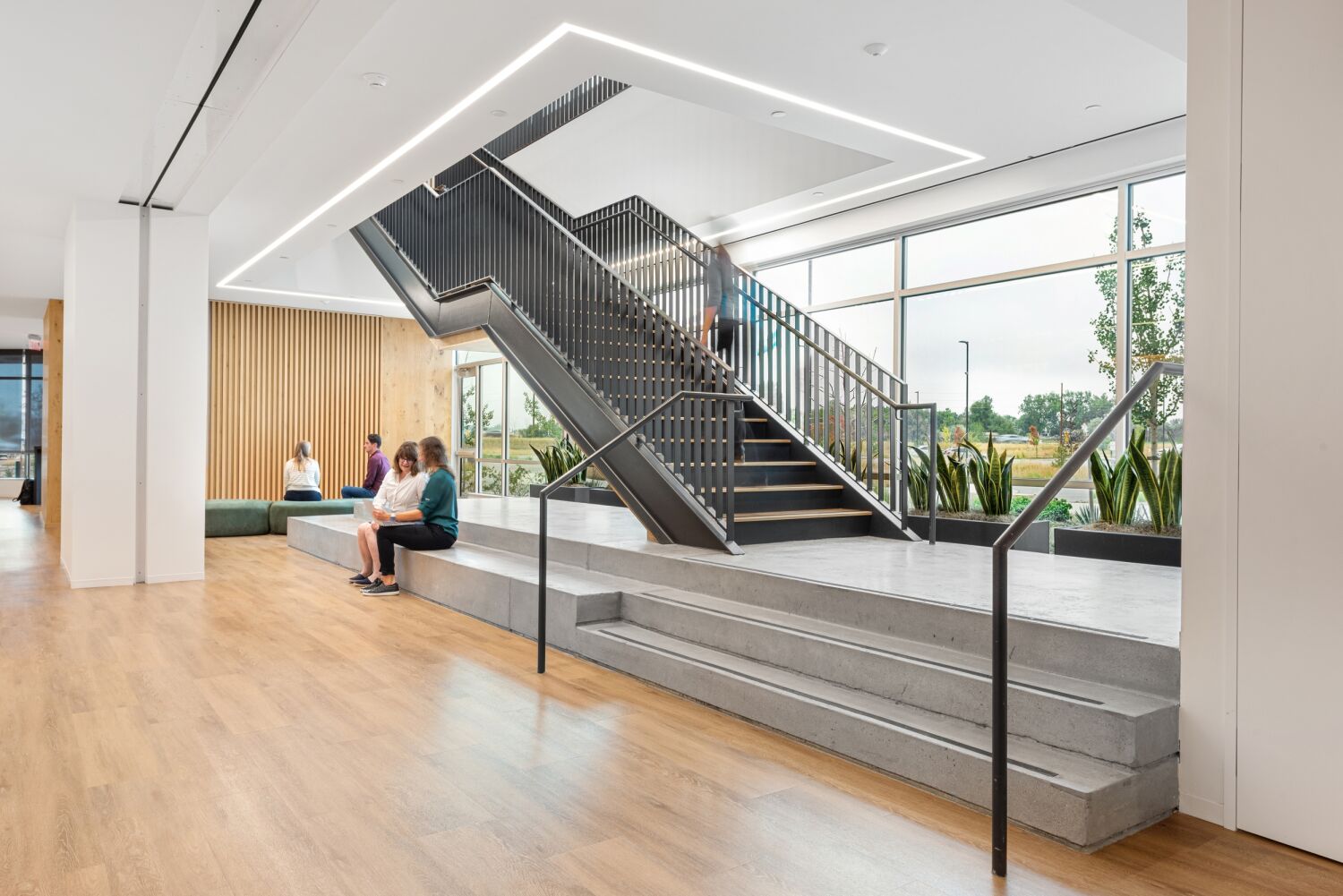 Interior workplace space at Medtronic featuring a large open staircase with metal railings, concrete tiered seating, wood flooring, floor-to-ceiling windows, and built-in bench seating along a slatted wood wall.
