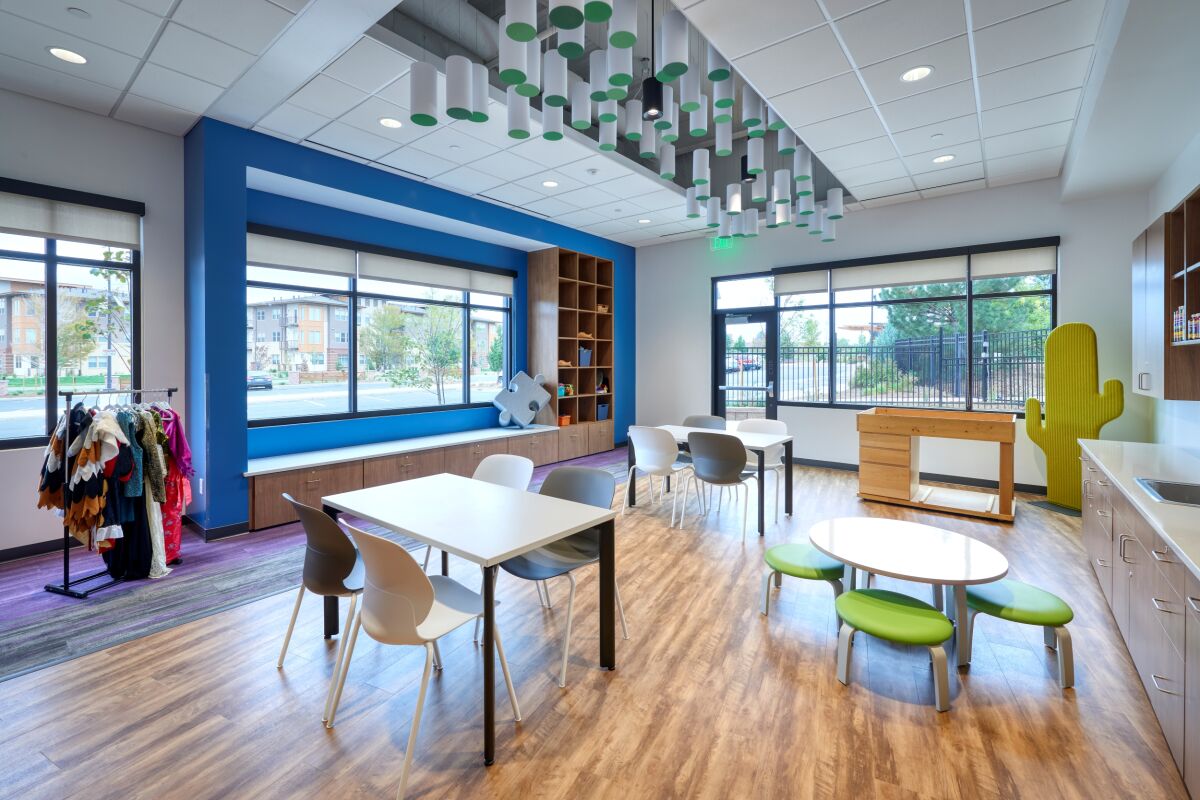 Family activity room at the Amy Davis Hospice Support Center featuring white tables with mixed chairs, a circular children’s table with green seats, built‑in shelving, large windows with views outside, wood‑look flooring, and a ceiling installation of hang