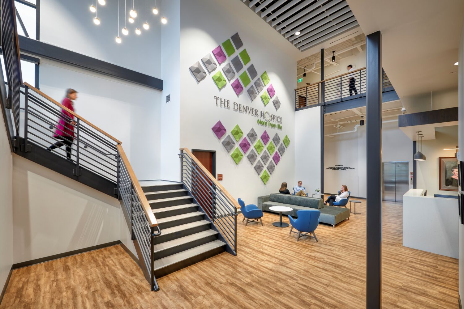Lobby gathering space at The Denver Hospice featuring a geometric feature wall with colorful panels, a large staircase with metal railings, pendant lighting, wood‑look flooring, tall windows, and contemporary lounge seating near the entrance.