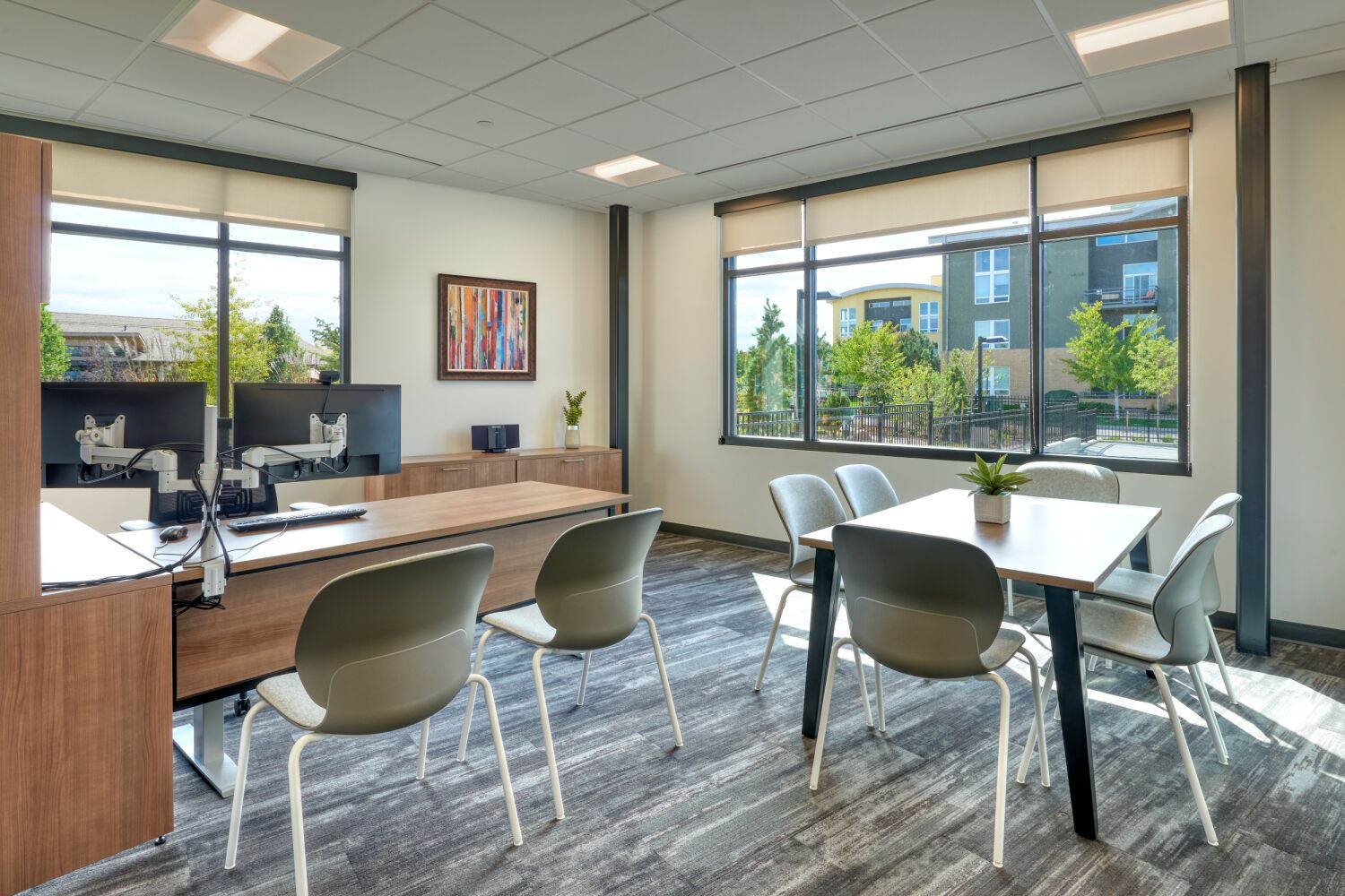 Private office at the Amy Davis Hospice Support Center featuring a wood desk with dual monitors, gray guest chairs, a rectangular meeting table with seating, framed artwork, large windows with exterior views, and patterned carpet flooring.