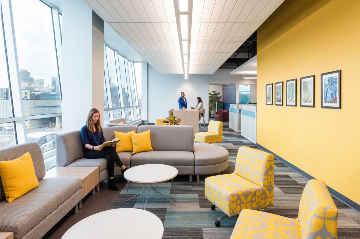 Lounge space in the Caring for Colorado Denver office featuring modern gray seating with yellow accent pillows, patterned yellow chairs, round white tables, a bright yellow feature wall with framed artwork, and large windows.