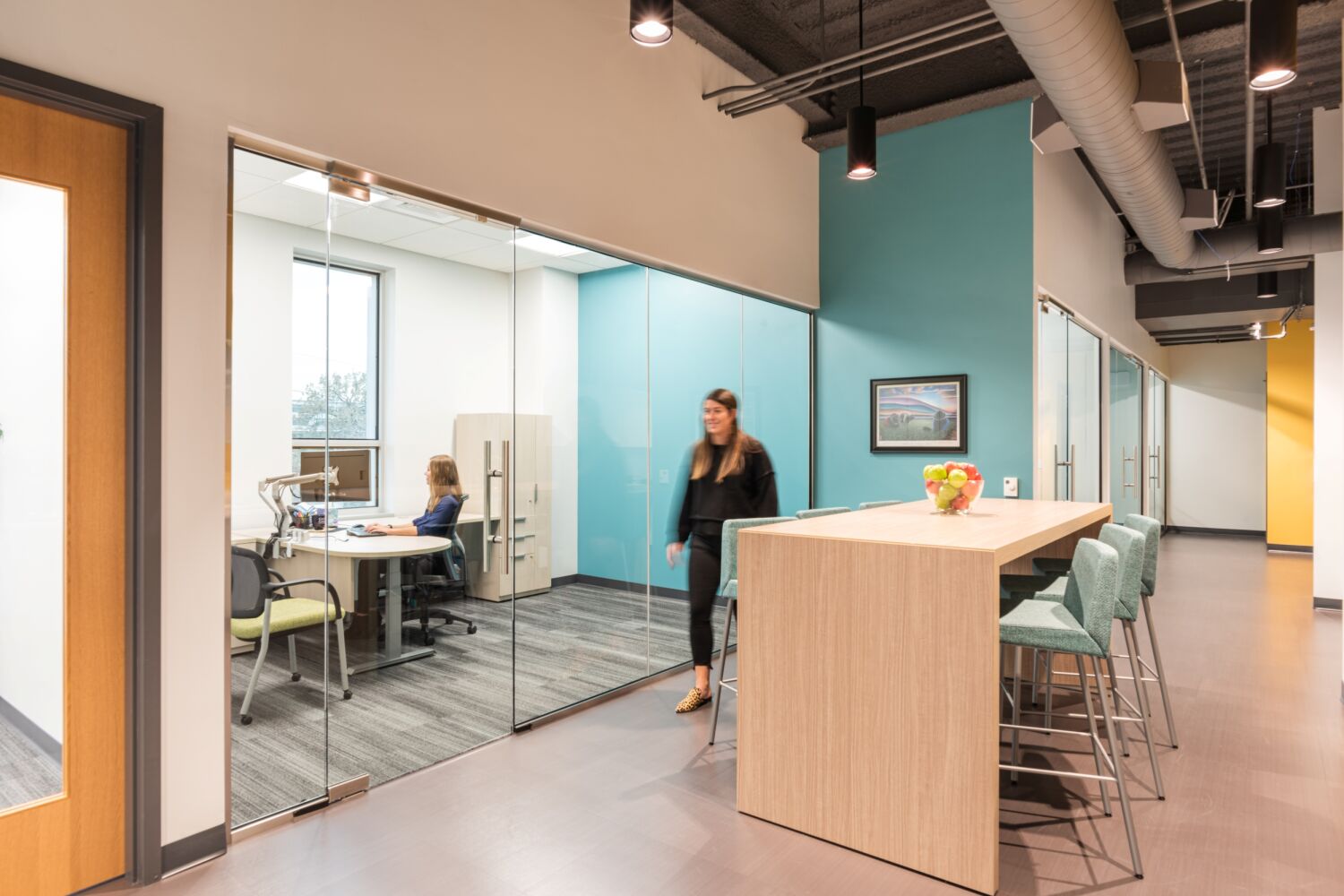 Hallway in the Caring for Colorado Denver office featuring glass-front meeting rooms, a tall wood collaboration table with upholstered stools, teal accent walls, exposed ceilings, and wood-look flooring.