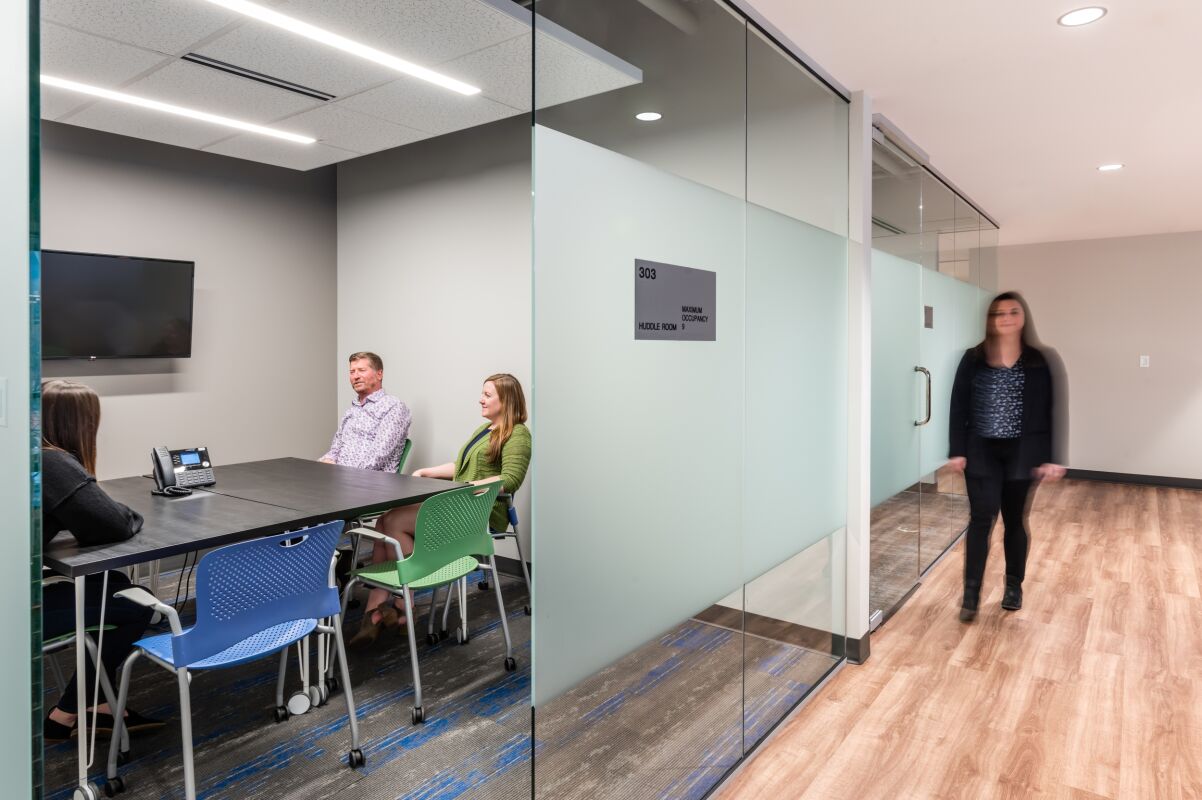 Glass‑front meeting room in the STRIDE Administration Office featuring a rectangular conference table with mixed chairs, a wall‑mounted display screen, carpet with blue accents, and a corridor with wood‑look flooring and overhead lighting.