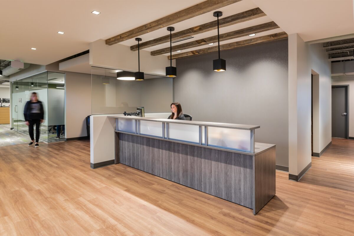 Reception area in the STRIDE Administration Office featuring a wood‑accented reception desk with frosted panels, pendant lighting with black fixtures, exposed ceiling beams, wood‑look flooring, and adjacent glass‑front office spaces.