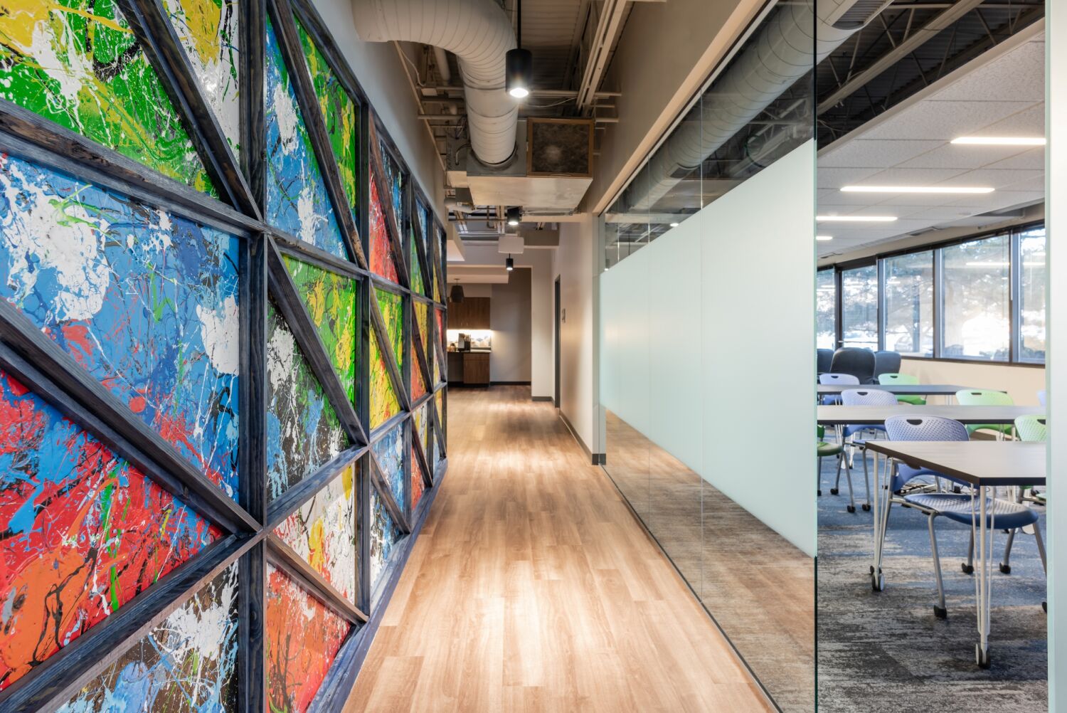 Hallway in the STRIDE Administration Office featuring a colorful geometric art installation along one wall, wood‑look flooring, exposed ceiling structure, and a glass partition revealing an adjacent training room with tables and chairs.