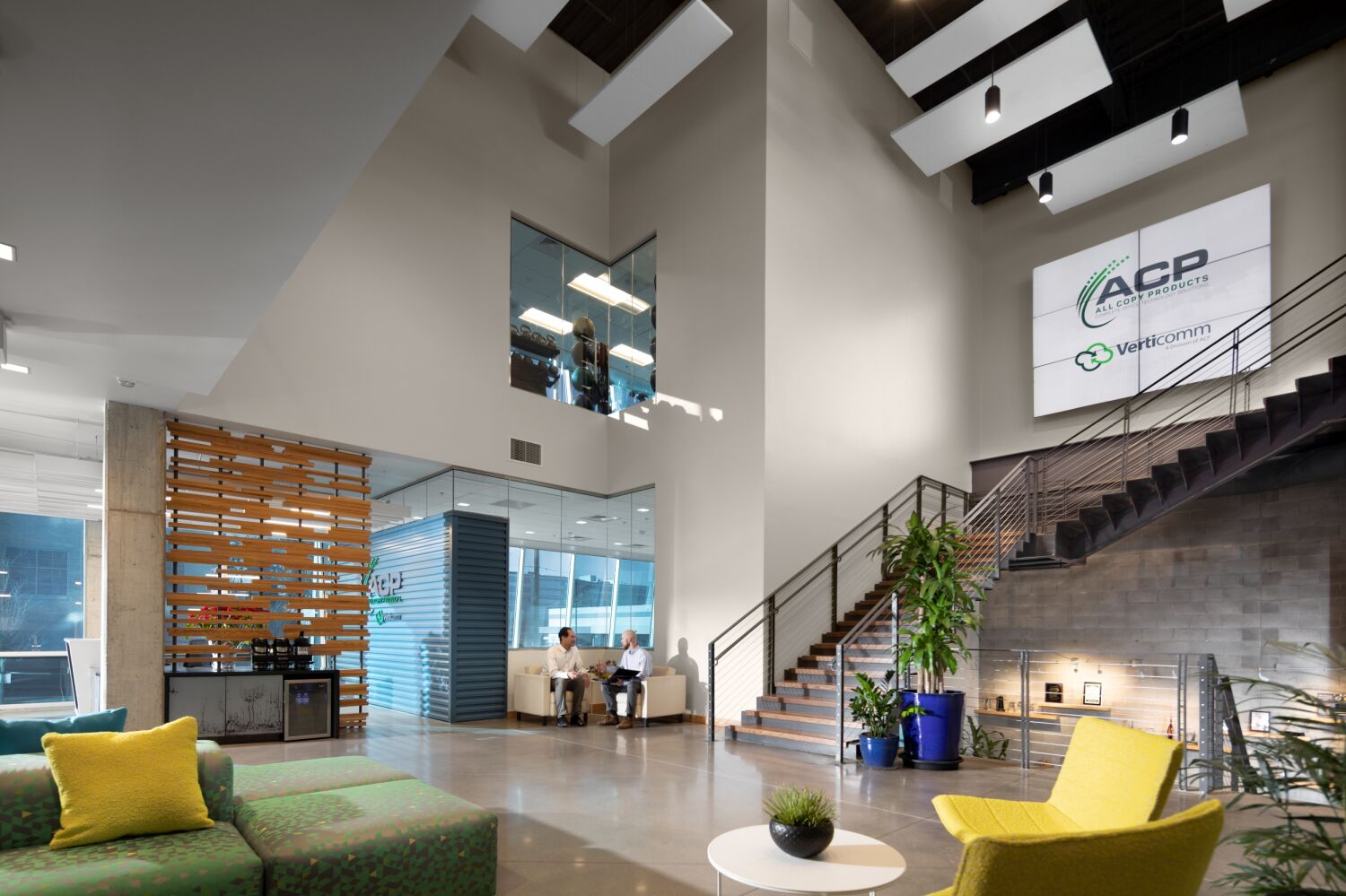 Atrium lobby at the All Copy Headquarters featuring a central staircase with metal railings, a tall feature wall with the ACP logo, polished concrete flooring, colorful lounge seating, suspended acoustic ceiling panels, and a wood‑slat partition near glass