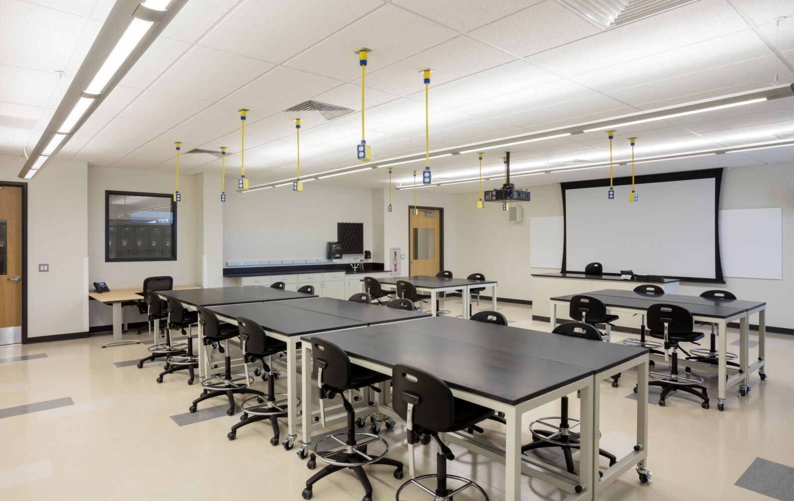Lab classroom with worktables, stools, overhead utility drops, and a projection screen.