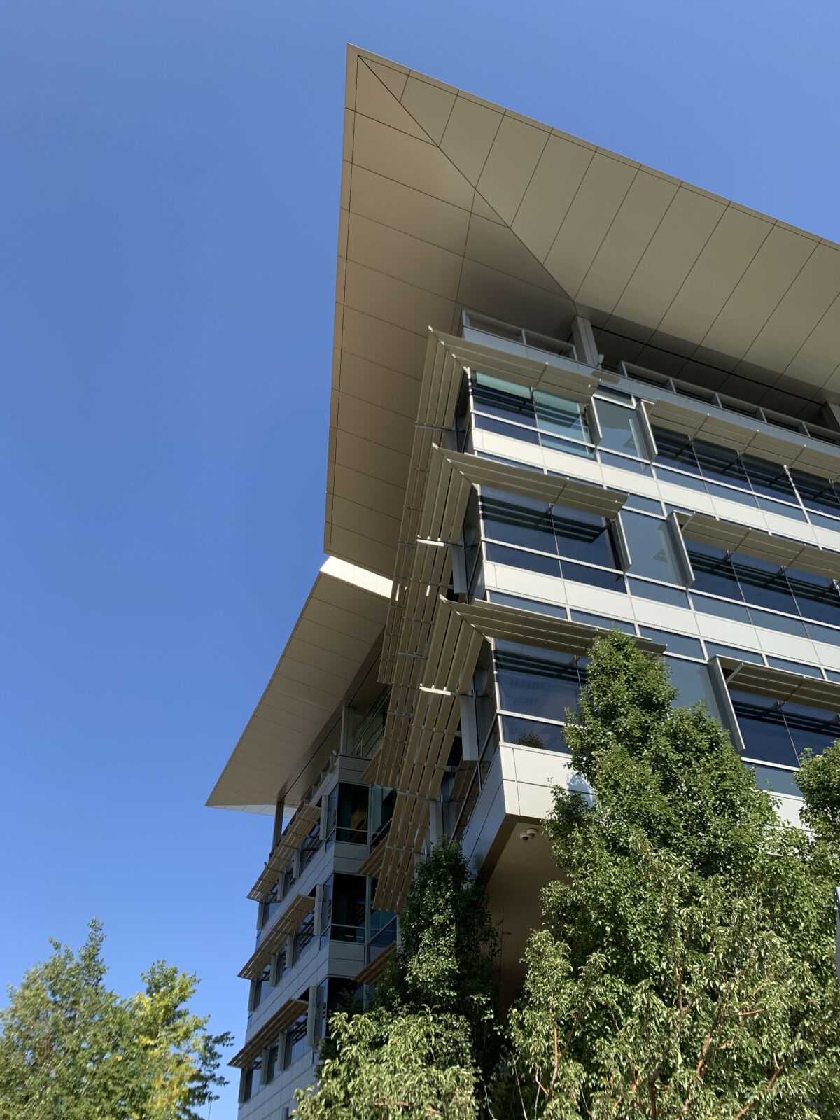 Upward angled view of a modern office building with large glass panels, layered sunshades, and an extended overhanging roof against a clear blue sky.