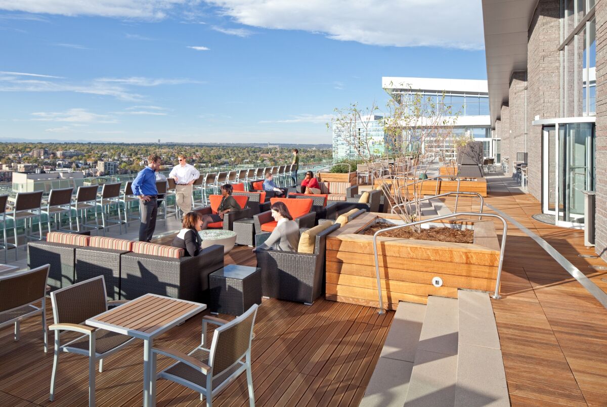 Rooftop amenity terrace at the DaVita Headquarters featuring outdoor lounge seating, modern planters, wood decking, dining tables with chairs, glass railing, and expansive city and mountain views.