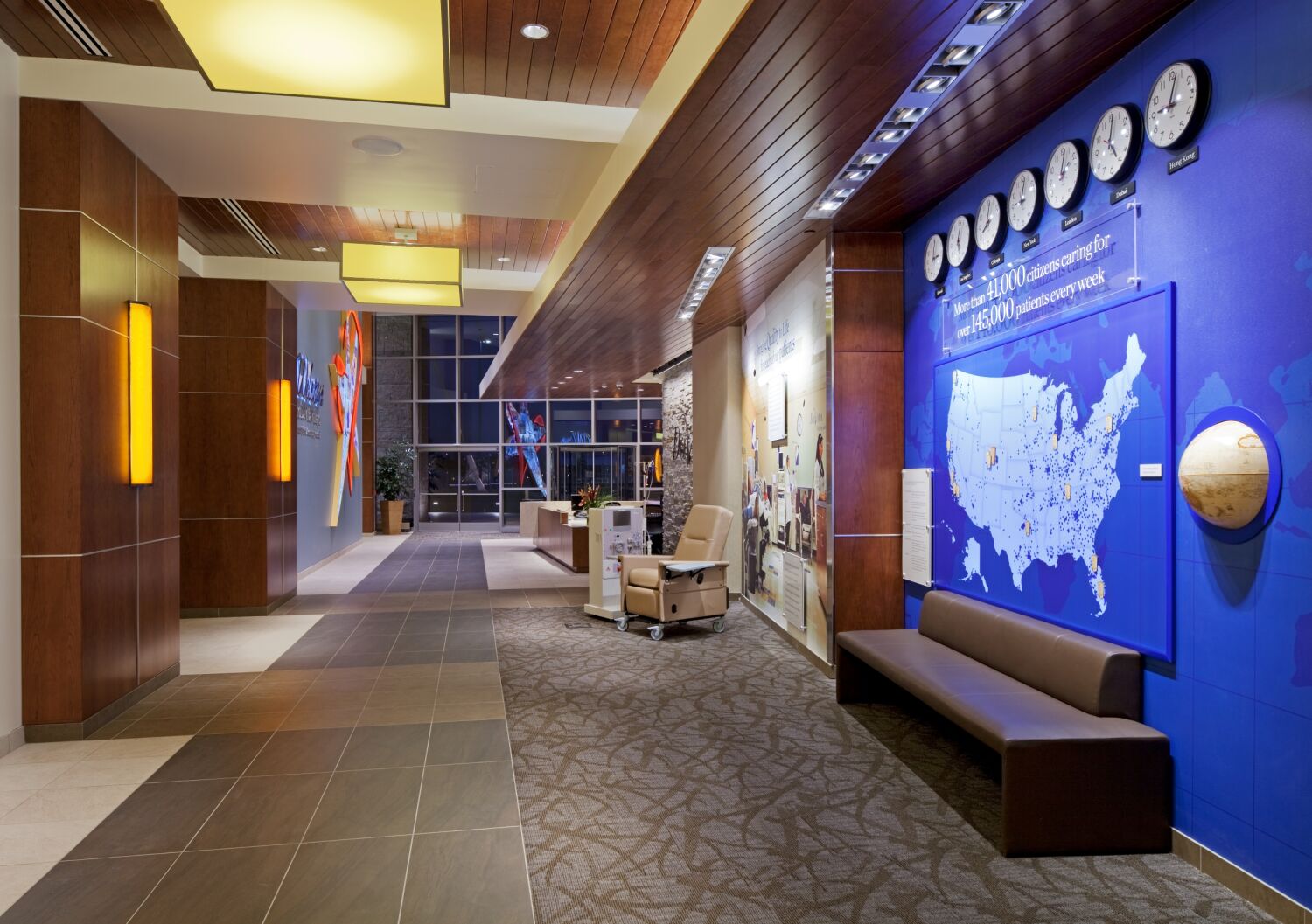 Interior corridor at the DaVita Headquarters featuring a blue branding wall with multiple clocks, a U.S. map graphic, a wood‑slat ceiling, pendant lighting, stone and wood accents, carpet and tile flooring, and a bench along the feature wall.