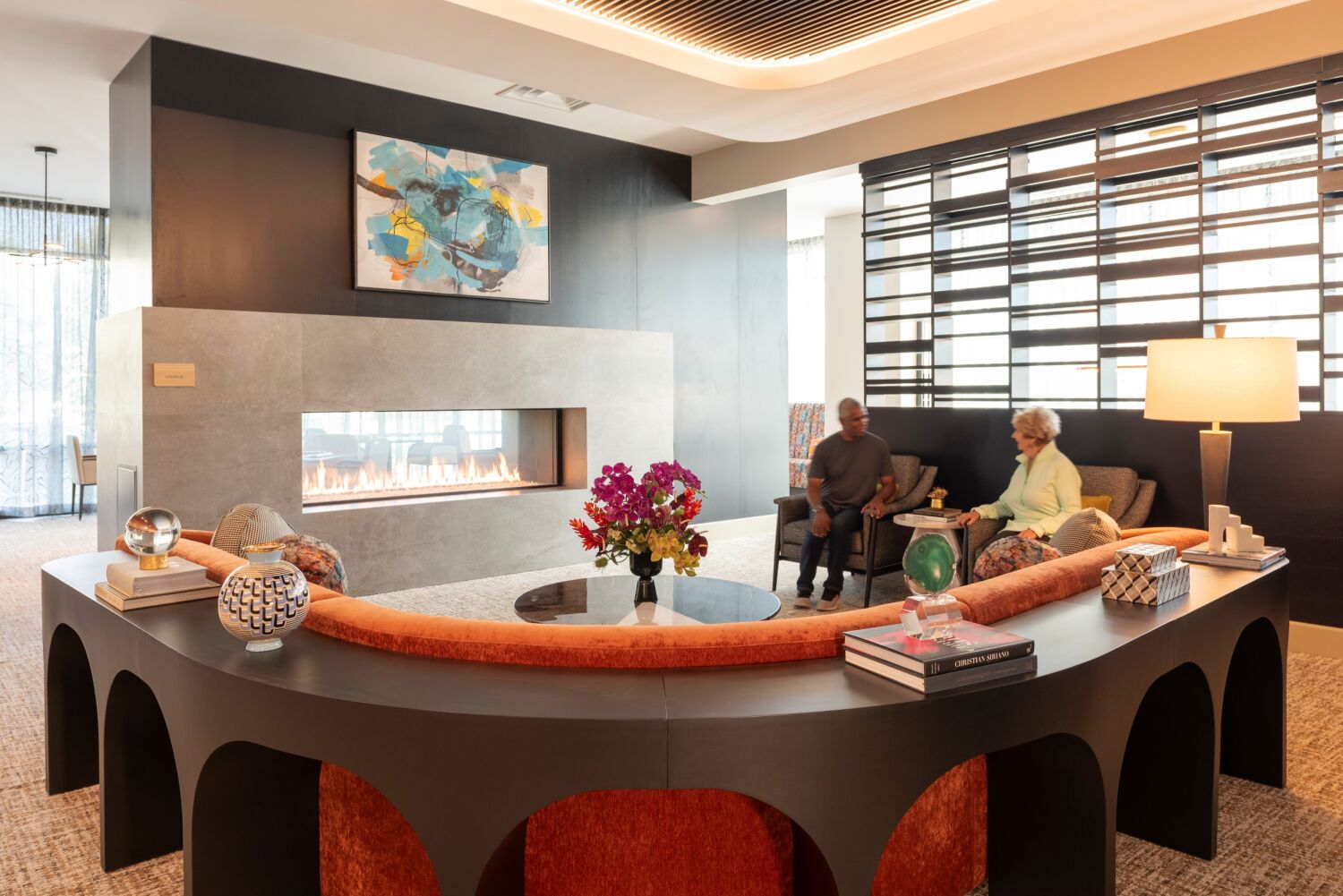 Lounge area with a curved upholstered sofa surrounding a modern linear fireplace, floral arrangement on a round coffee table, and seating arranged near a wall with horizontal slatted panels.