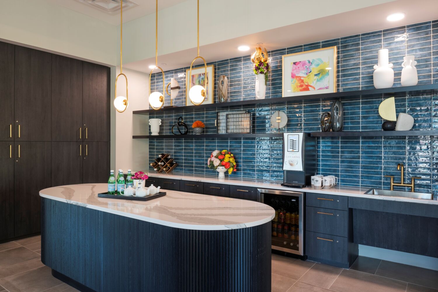 Modern café area with a curved marble‑top island, dark wood cabinetry, blue tile backsplash, open shelving with decorative items, pendant lighting, and an under‑counter beverage refrigerator.