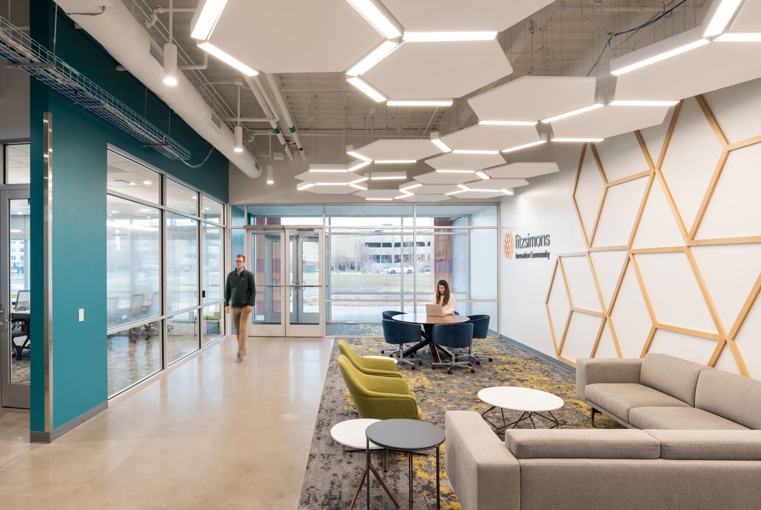 Lobby area with geometric ceiling lights, teal accent wall, glass meeting rooms, and lounge seating near the entrance.
