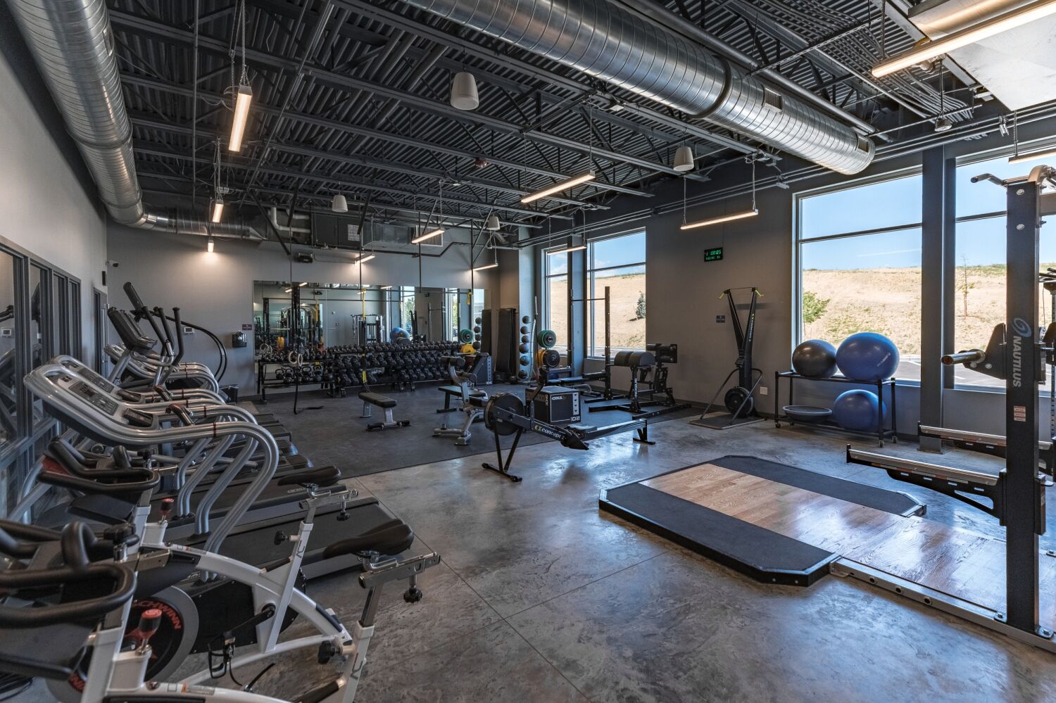 Strength training room with cardio machines, free weights, benches, and large windows providing natural light.