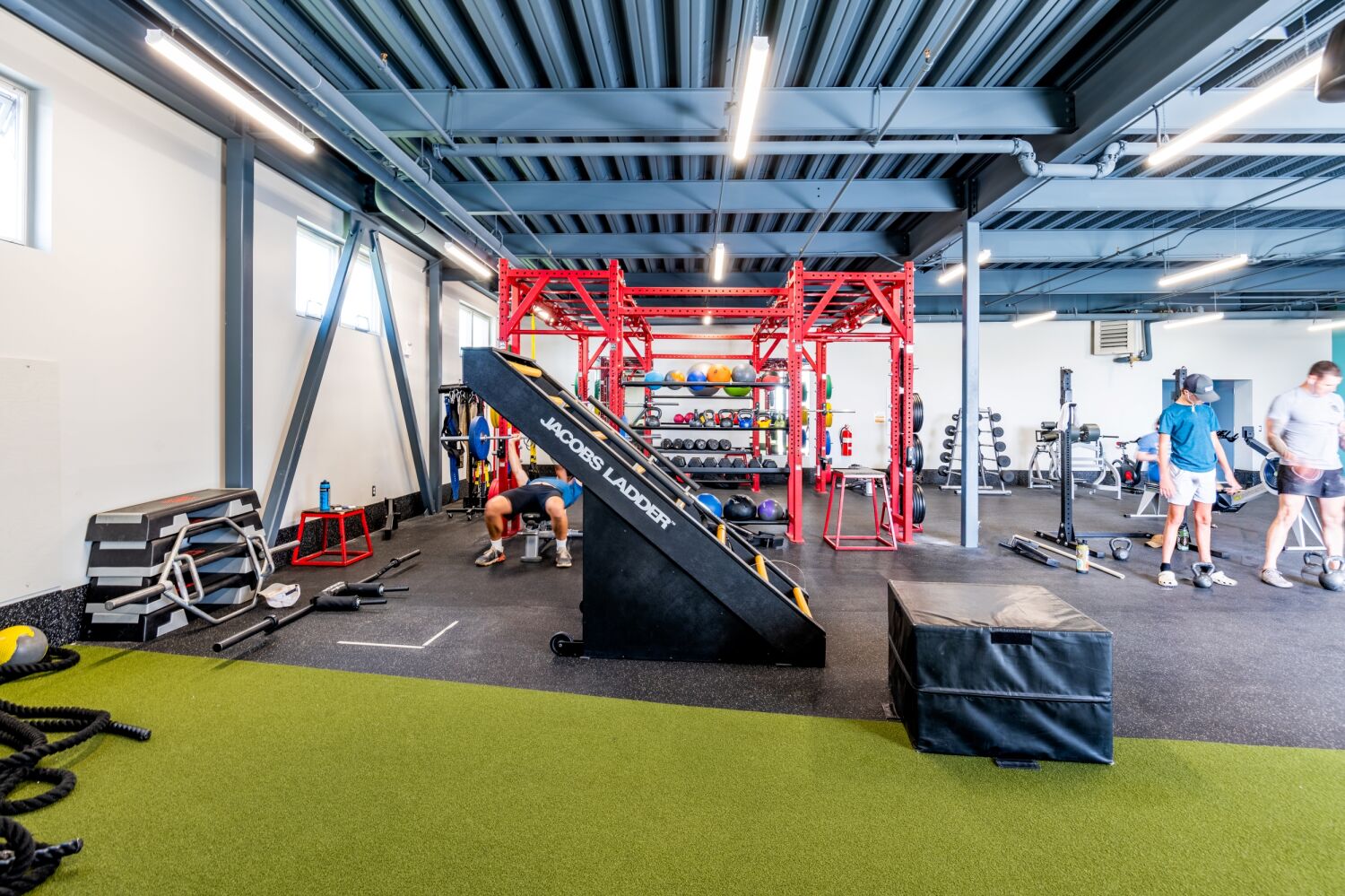 Functional fitness area with a Jacobs Ladder machine, red training rig, free weights, turf flooring, and an open exposed‑ceiling structure.