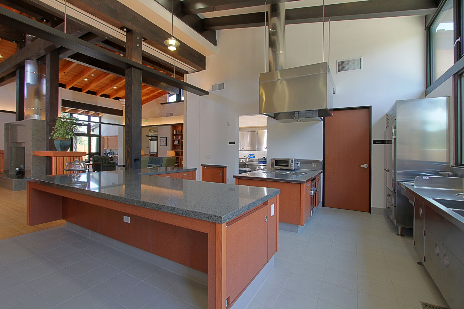 Community kitchen with large central island, stainless‑steel appliances, overhead ventilation hoods, wood‑beam ceiling, and adjacent open gathering spaces.