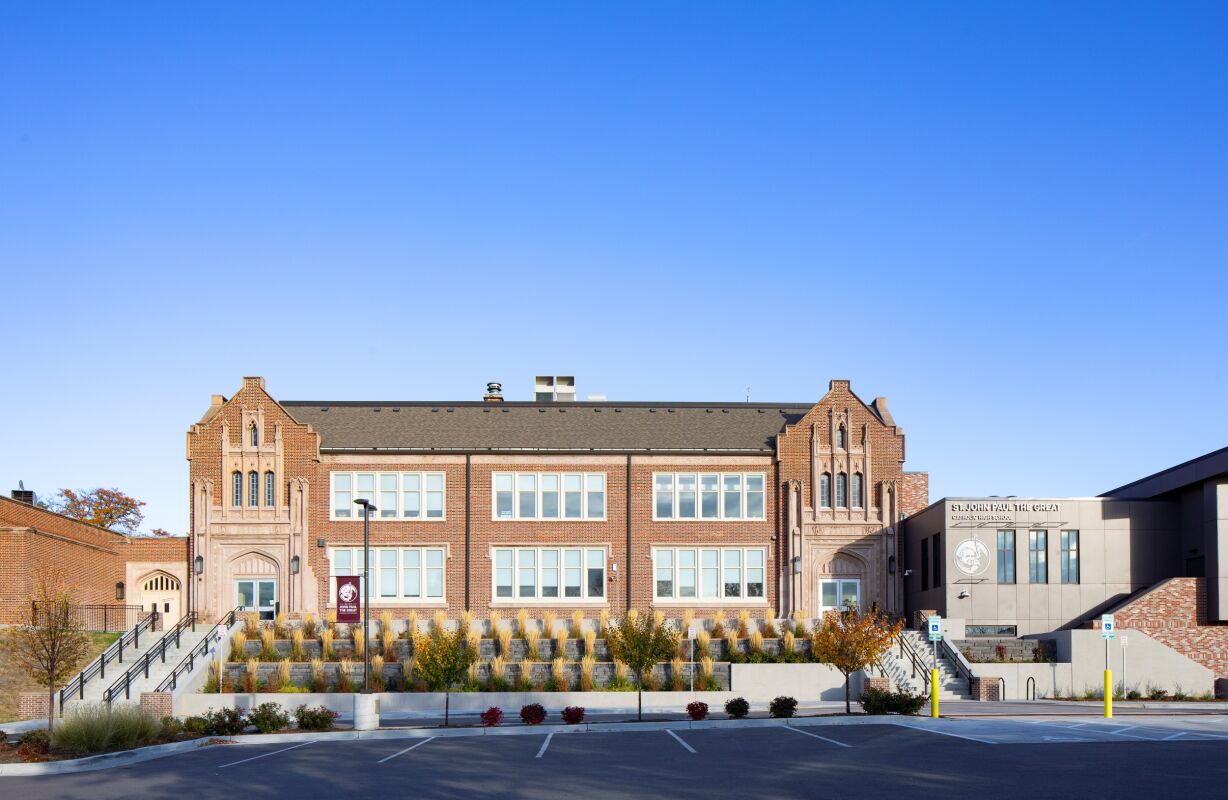 Front view of a historic brick school building with symmetrical façades, large multi‑pane windows, landscaped terraces, and an adjacent modern addition under a clear blue sky.