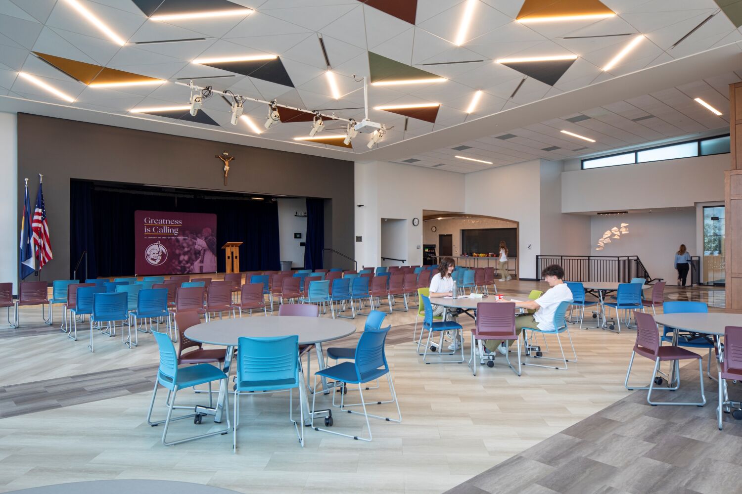 Commons space with patterned acoustic ceiling panels, round and rectangular tables, colorful chairs, and a stage with a podium and backdrop featuring the school emblem.
