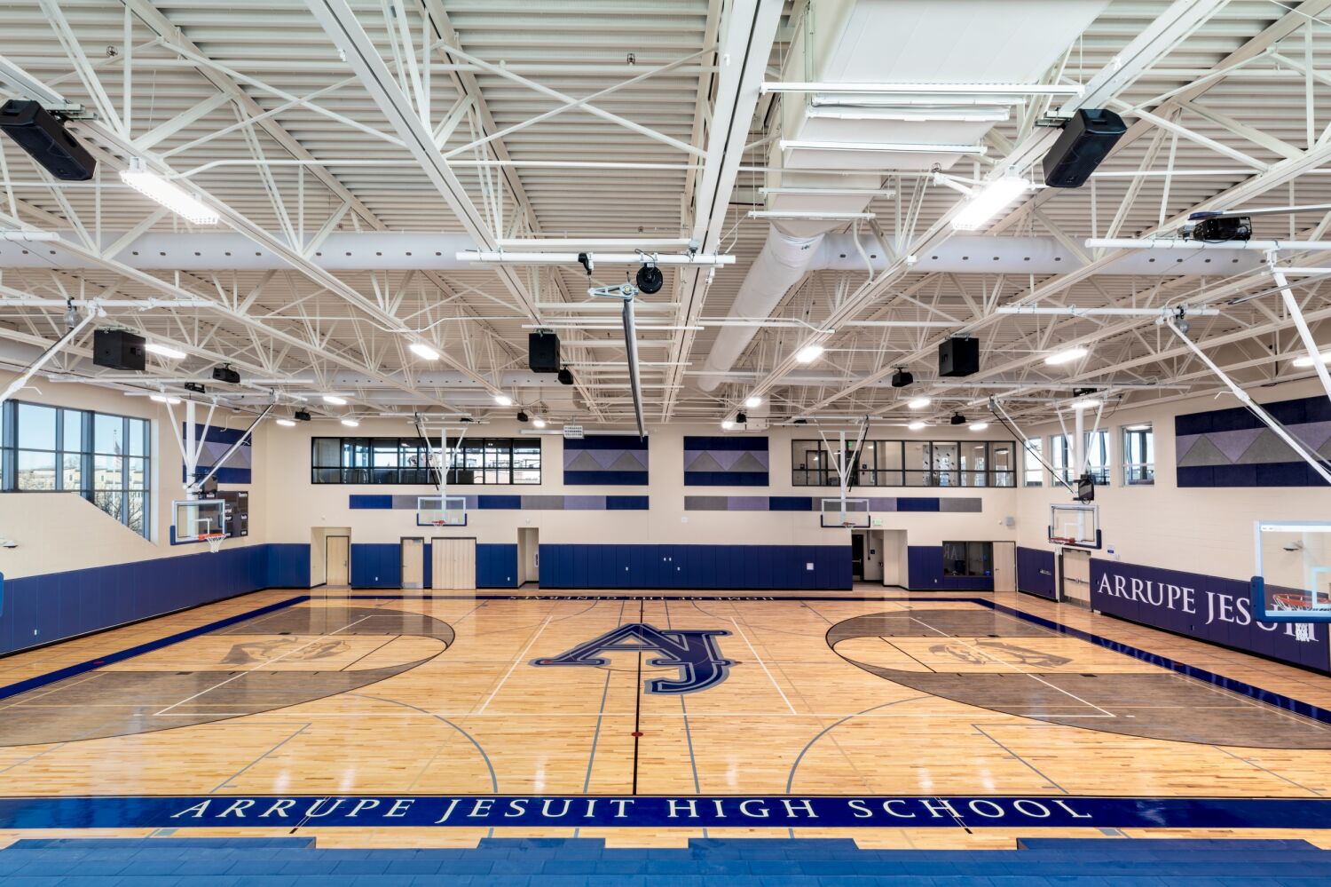 High school gymnasium with a full basketball court, wood flooring featuring a large school logo at center court, blue and gray acoustic wall panels, multiple backboards, and an exposed structural ceiling with suspended lighting.