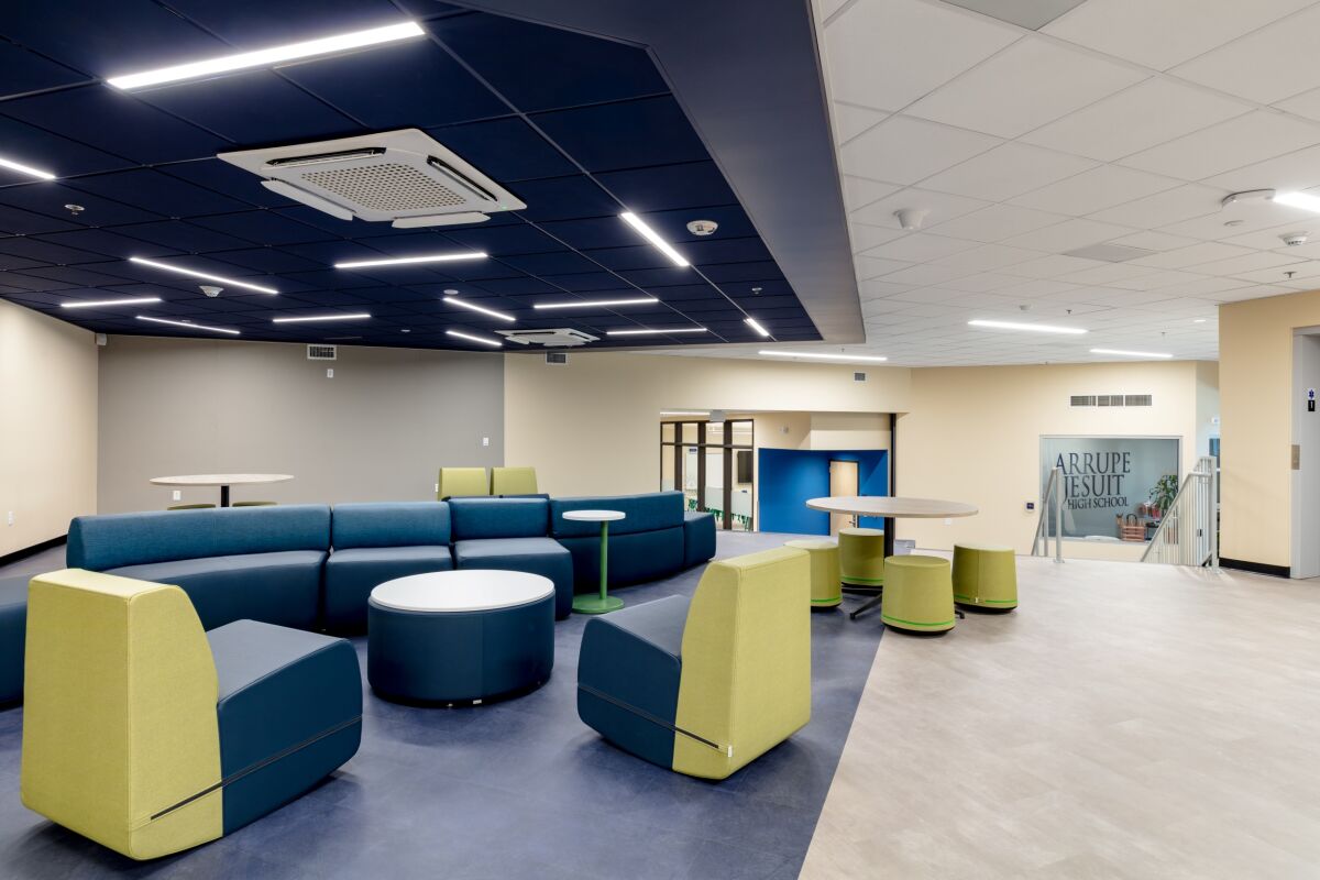 Student commons area with modular lounge seating in blue and green tones, circular tables, wood‑slat ceiling panels with integrated linear lighting, and adjacent glass‑front entry area.
