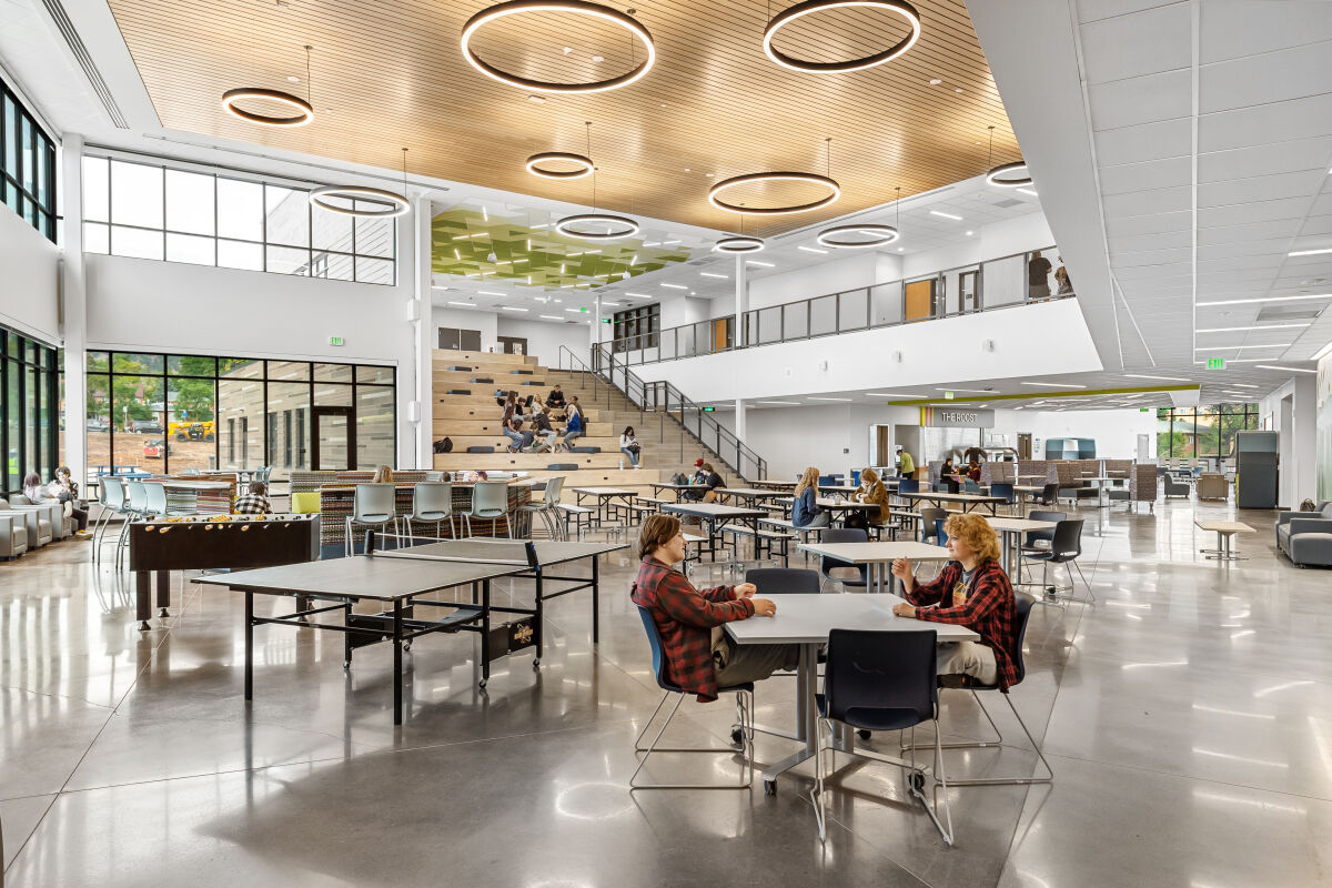 Open two‑story commons, floor‑to‑ceiling windows, cafeteria tables, a tiered learning stair with integrated cushions, and geometric green acoustic ceiling panels.