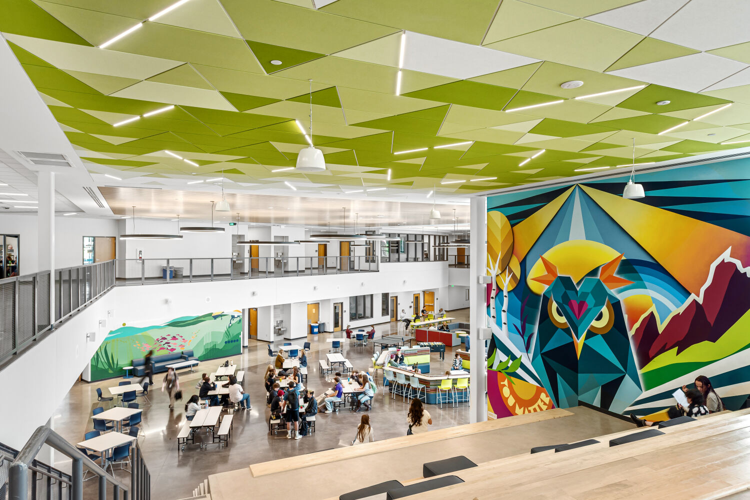 Two‑story school commons with polished concrete flooring, large windows, geometric green ceiling panels, cafeteria tables, curved lounge seating, wood‑slat ceiling accents, and a large colorful mural featuring a stylized owl.