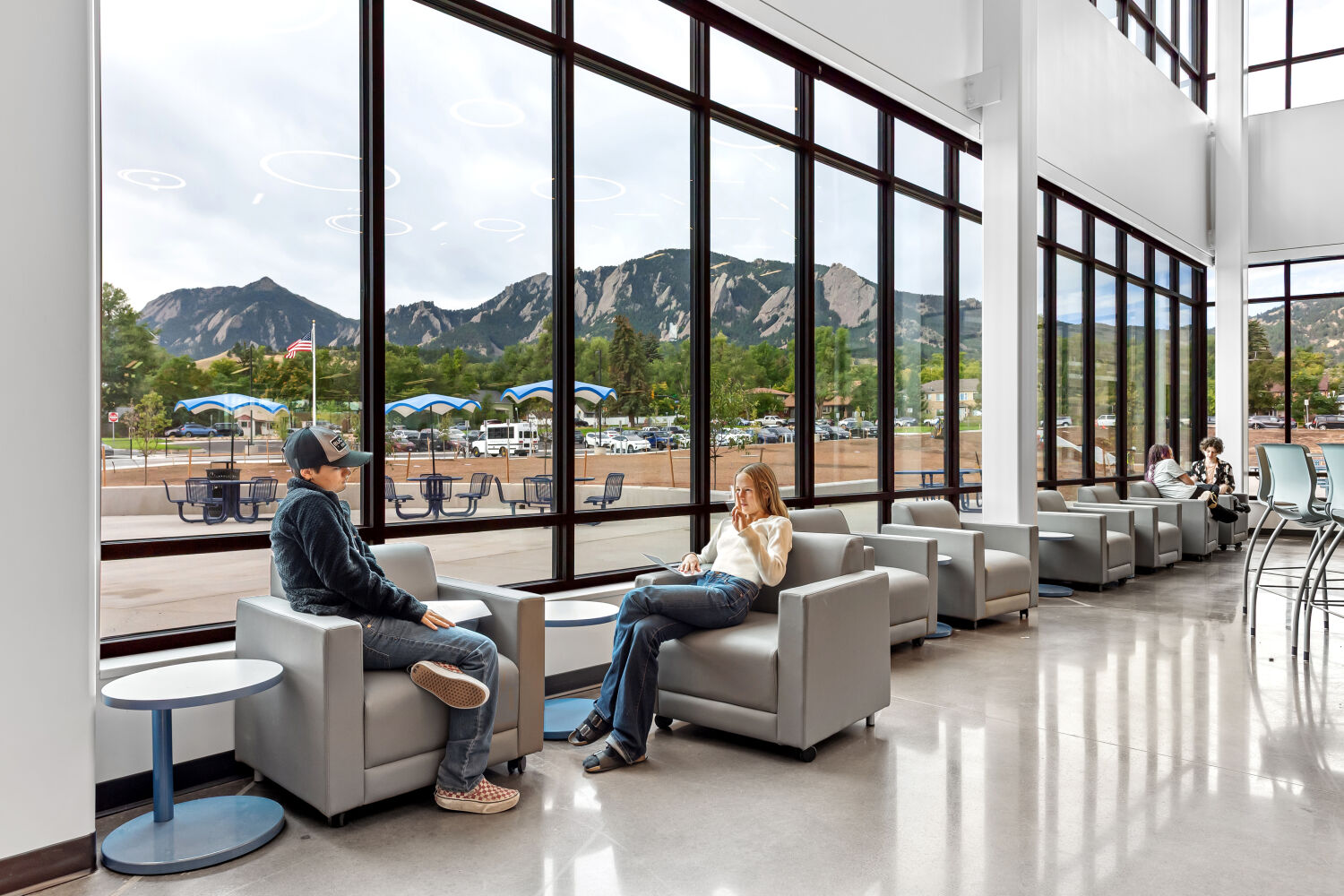 Lounge area with individual armchairs and small round side tables arranged along floor‑to‑ceiling windows, featuring polished concrete flooring, expansive mountain views, and high ceilings with suspended circular light fixtures.